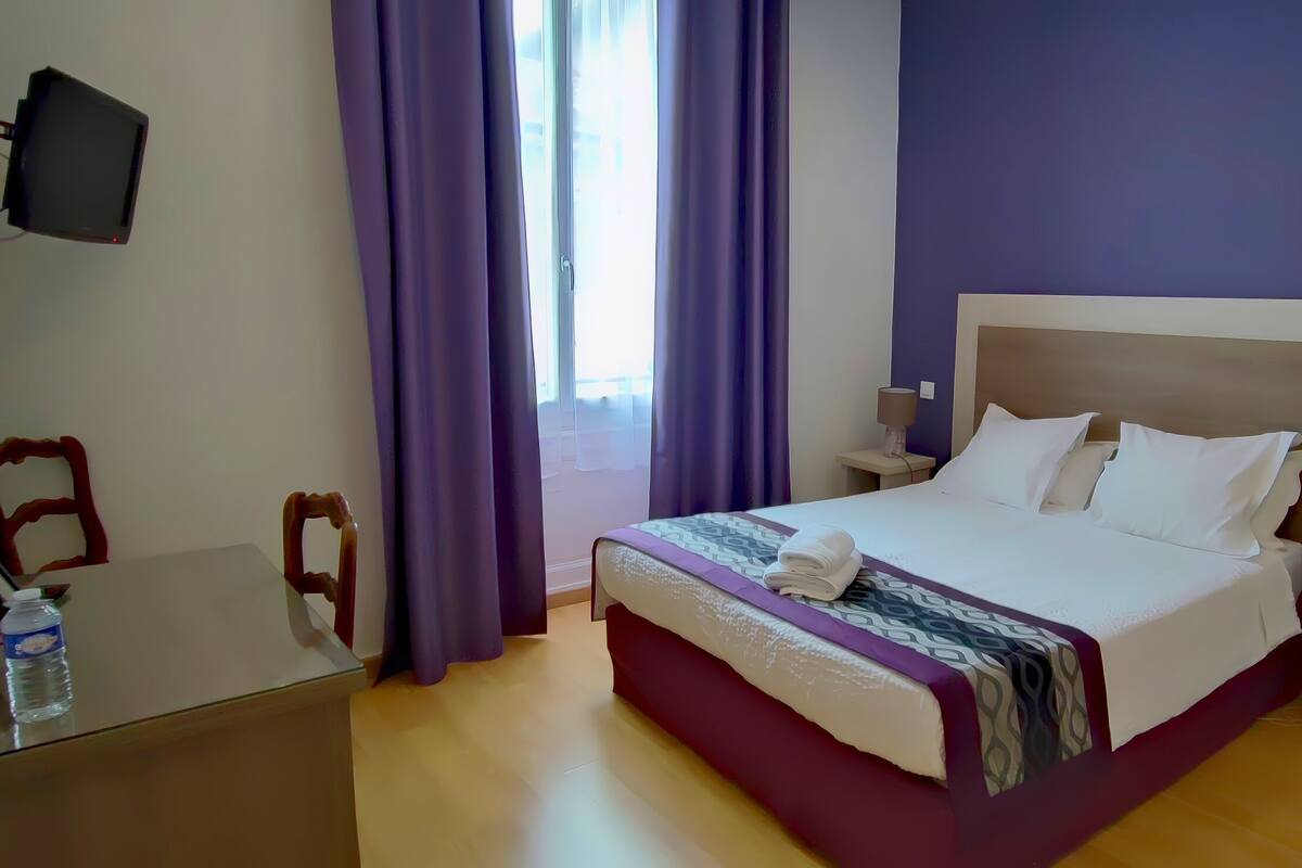 Une chambre d’hôtel avec un lit, une table de chevet, une lampe, un téléviseur fixé au mur et une fenêtre avec des rideaux violets.