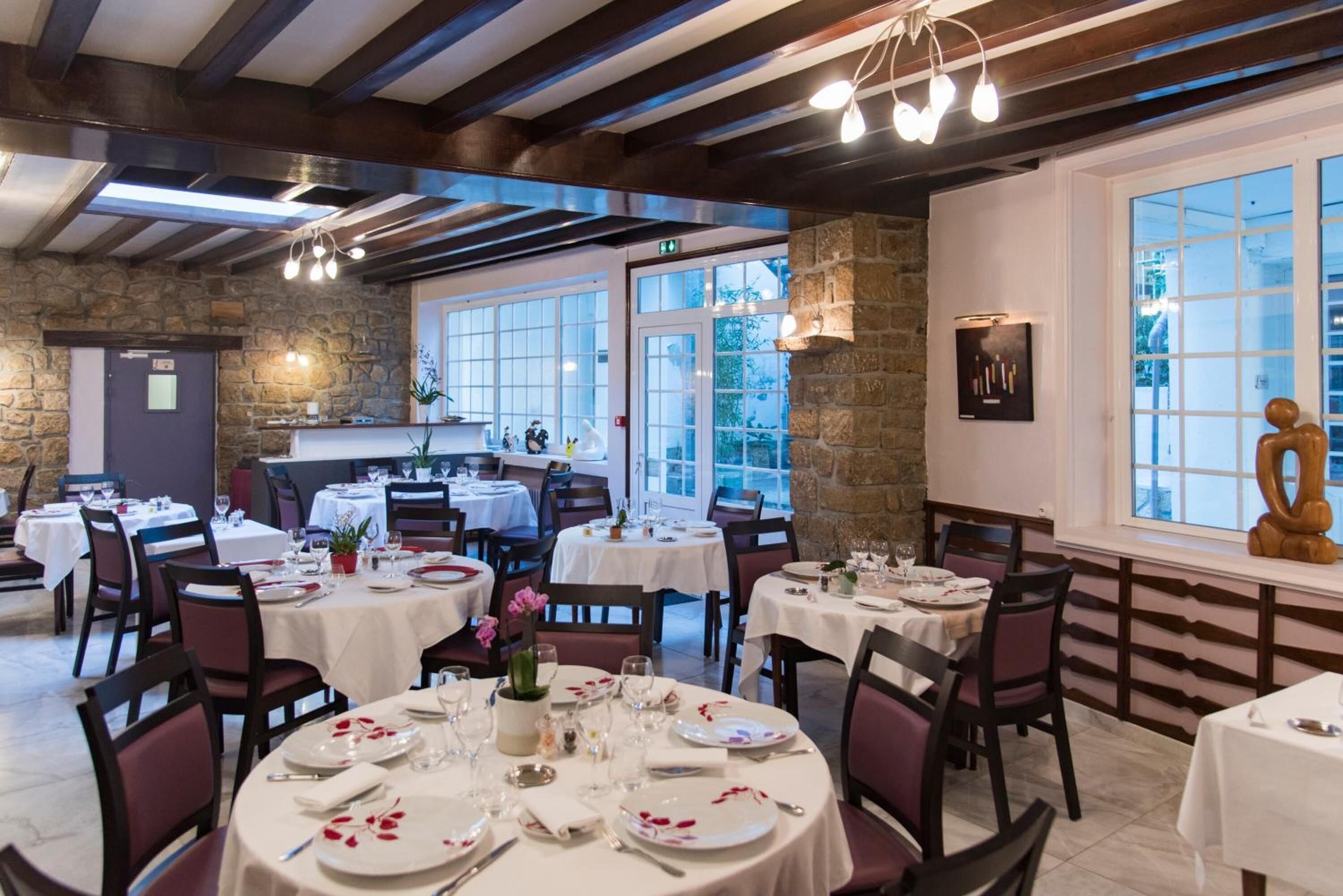 Un restaurante con mesas redondas dispuestas para comer, paredes de piedra y ventanas grandes.