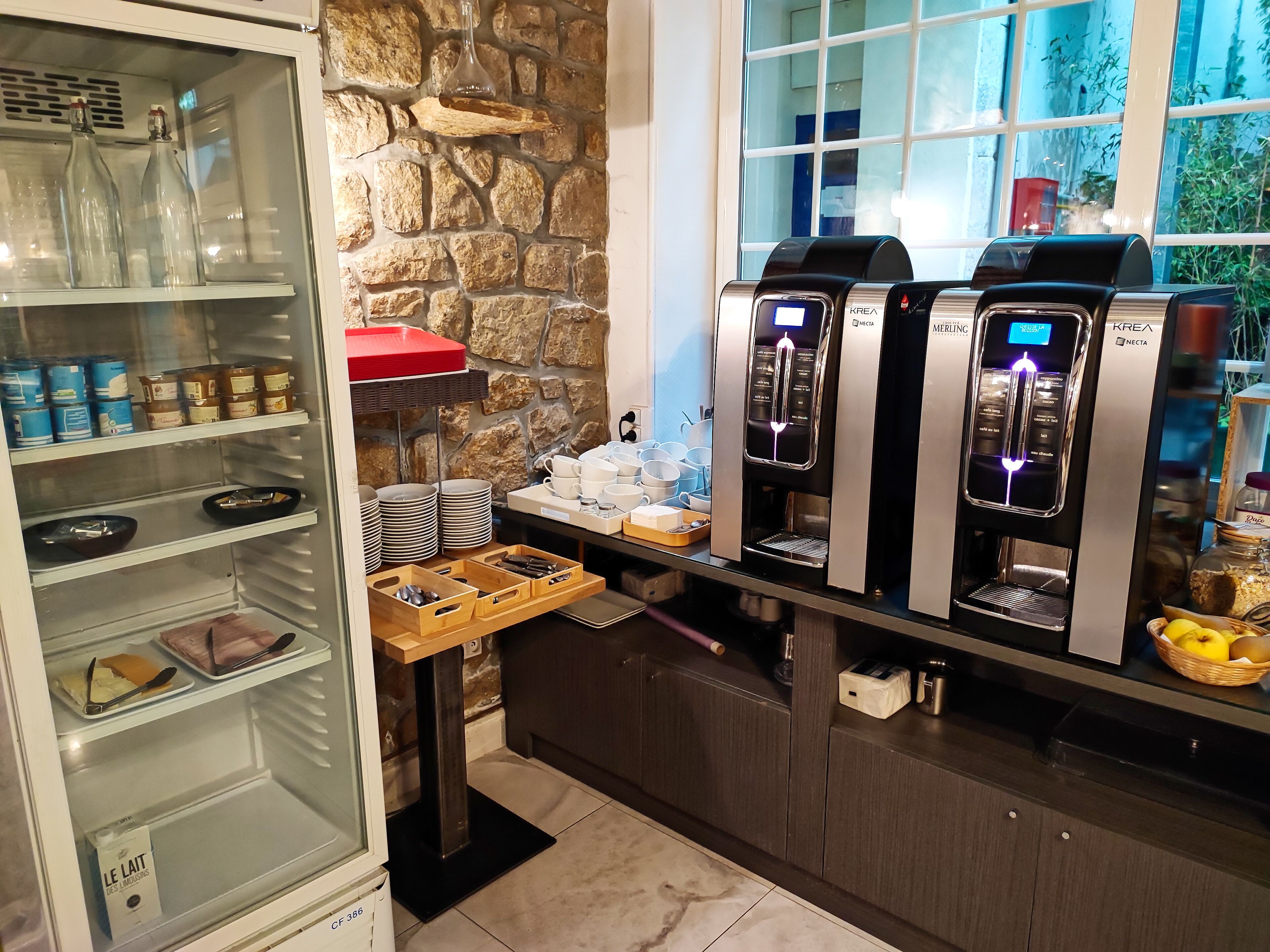 A kitchen area with a stone wall, a coffee station with KREA branded machines, and a glass-fronted refrigerator stocked with various items.