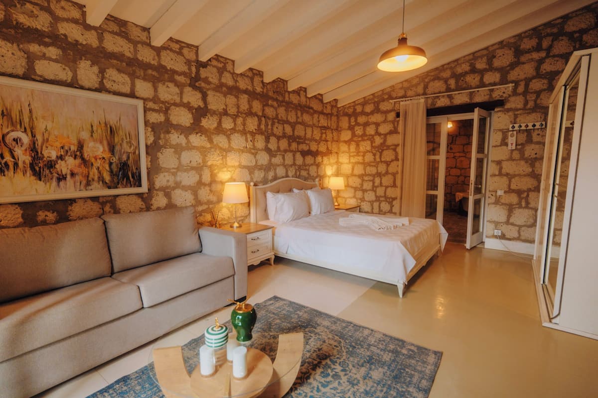 A bedroom with a stone wall, a bed with white linens, a sofa, a nightstand with lamps, and a painting on the wall.