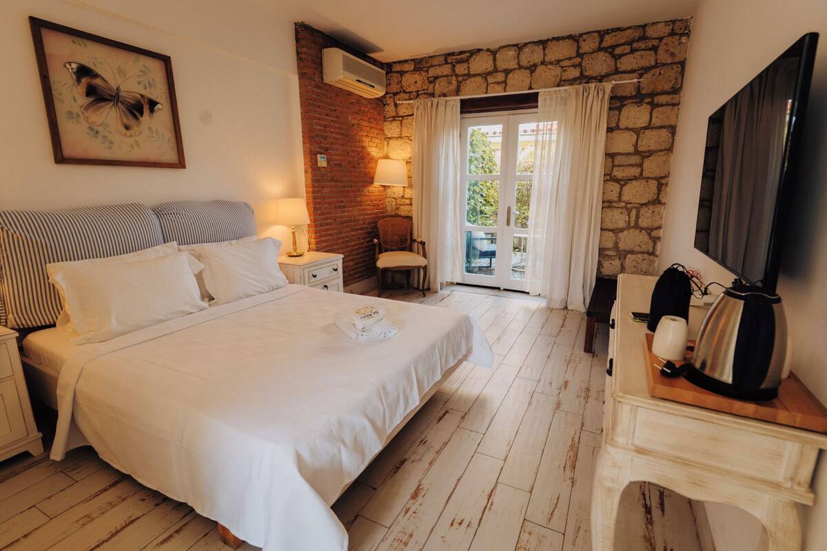 A hotel room with a large bed, a bedside table with a lamp, a chair, a stone wall, a window with curtains, and a flat-screen TV.