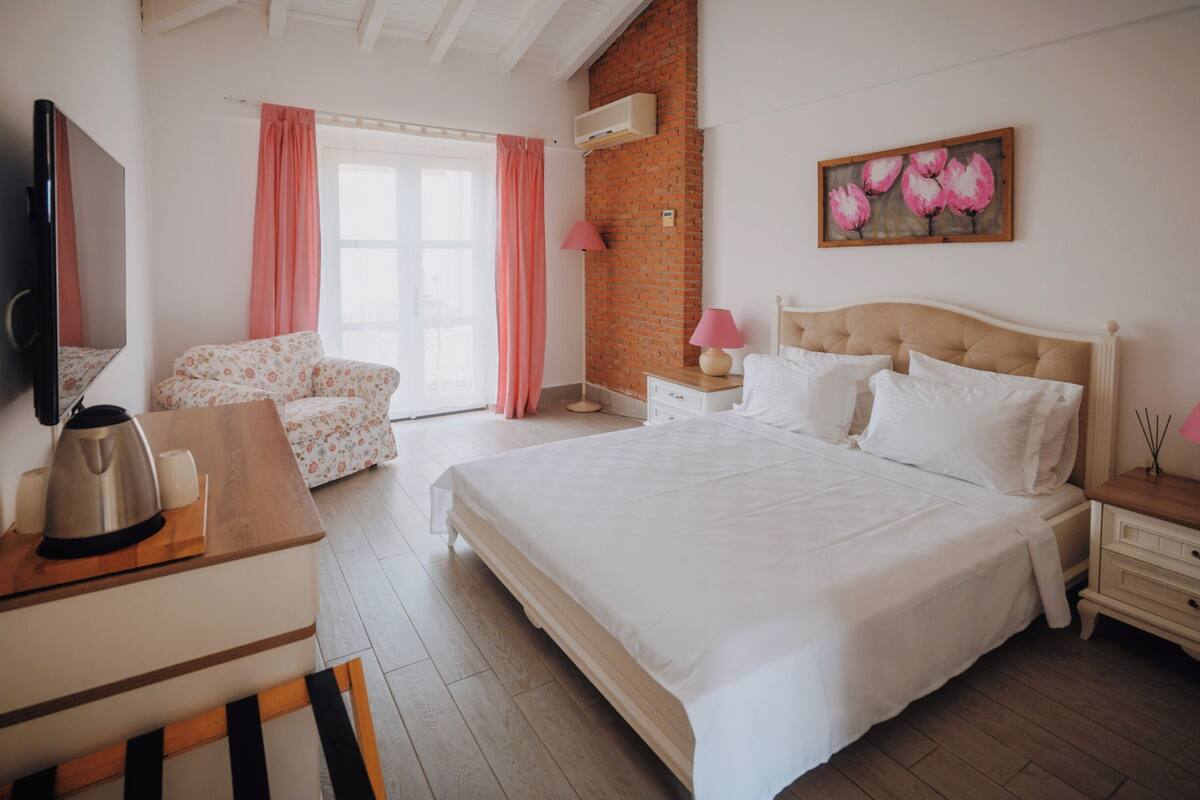A bedroom with a large bed, a desk with a kettle, a chair, a nightstand, and a wall-mounted TV.