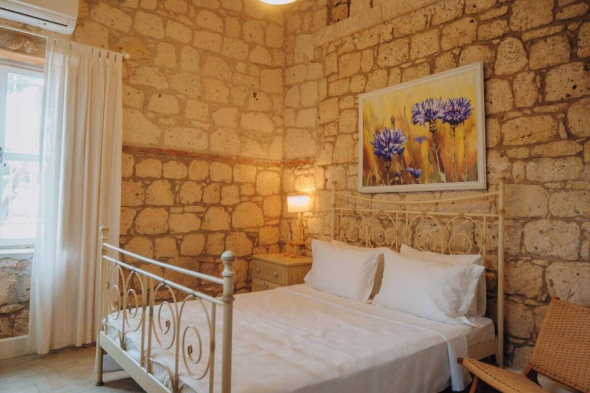 A bedroom with a stone wall, a bed with white bedding, a hanging light fixture, and a painting of flowers on the wall.
