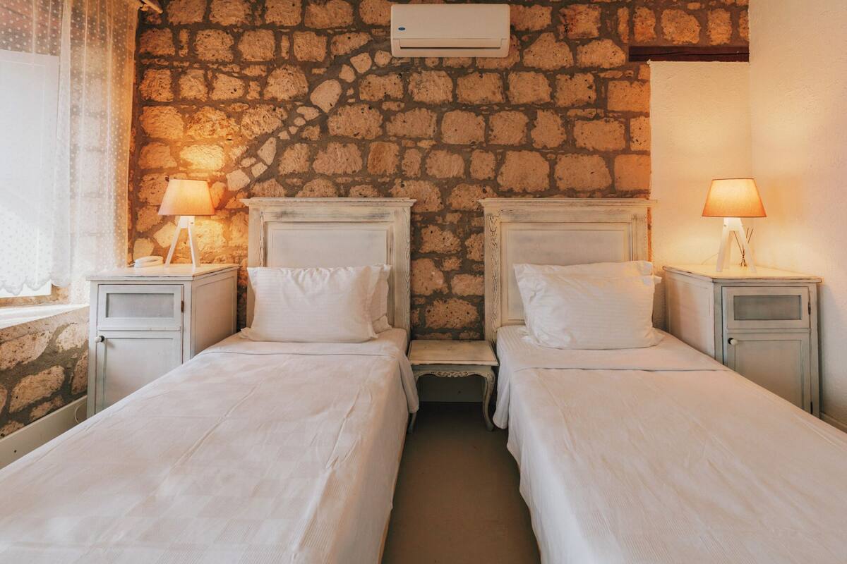 A room with two beds, each with a nightstand and a lamp, set against a stone wall with an air conditioning unit above.