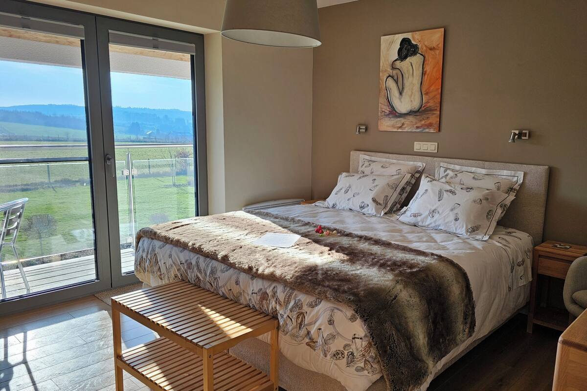 Een slaapkamer met een groot raam, een bed met opvallende kussens, een houten nachtkastje en een tafeltje met een decoratief object.