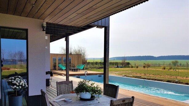 Una terraza de madera con piscina, mobiliario exterior y vistas al campo.