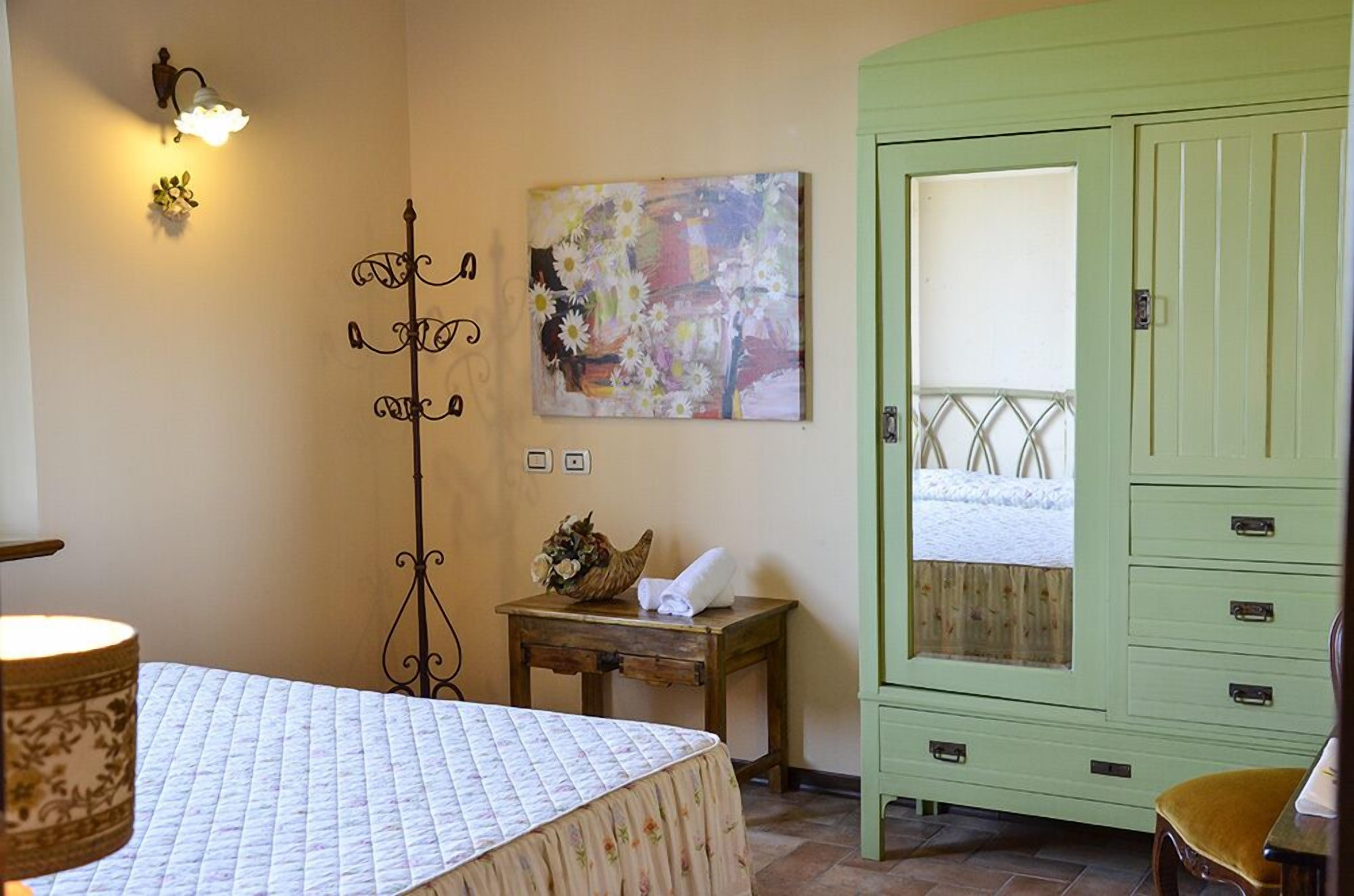 A room with a bed, a green wardrobe, a wall-mounted light fixture, a small table with a vase, and a painting.