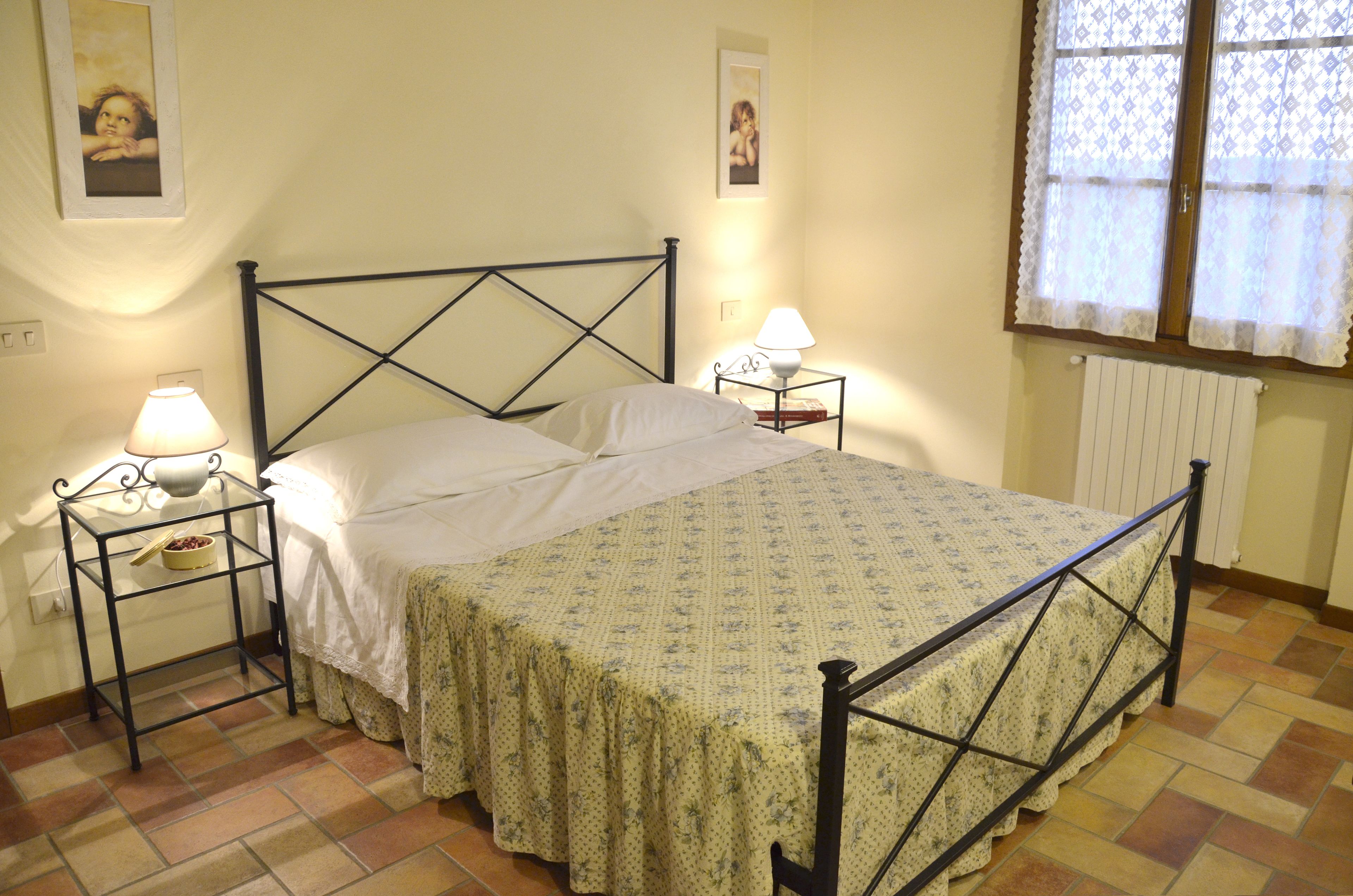 Una cama con estructura metálica, dos mesitas de noche con lámparas, una colcha estampada con motivos florales y una ventana con cortinas de encaje.
