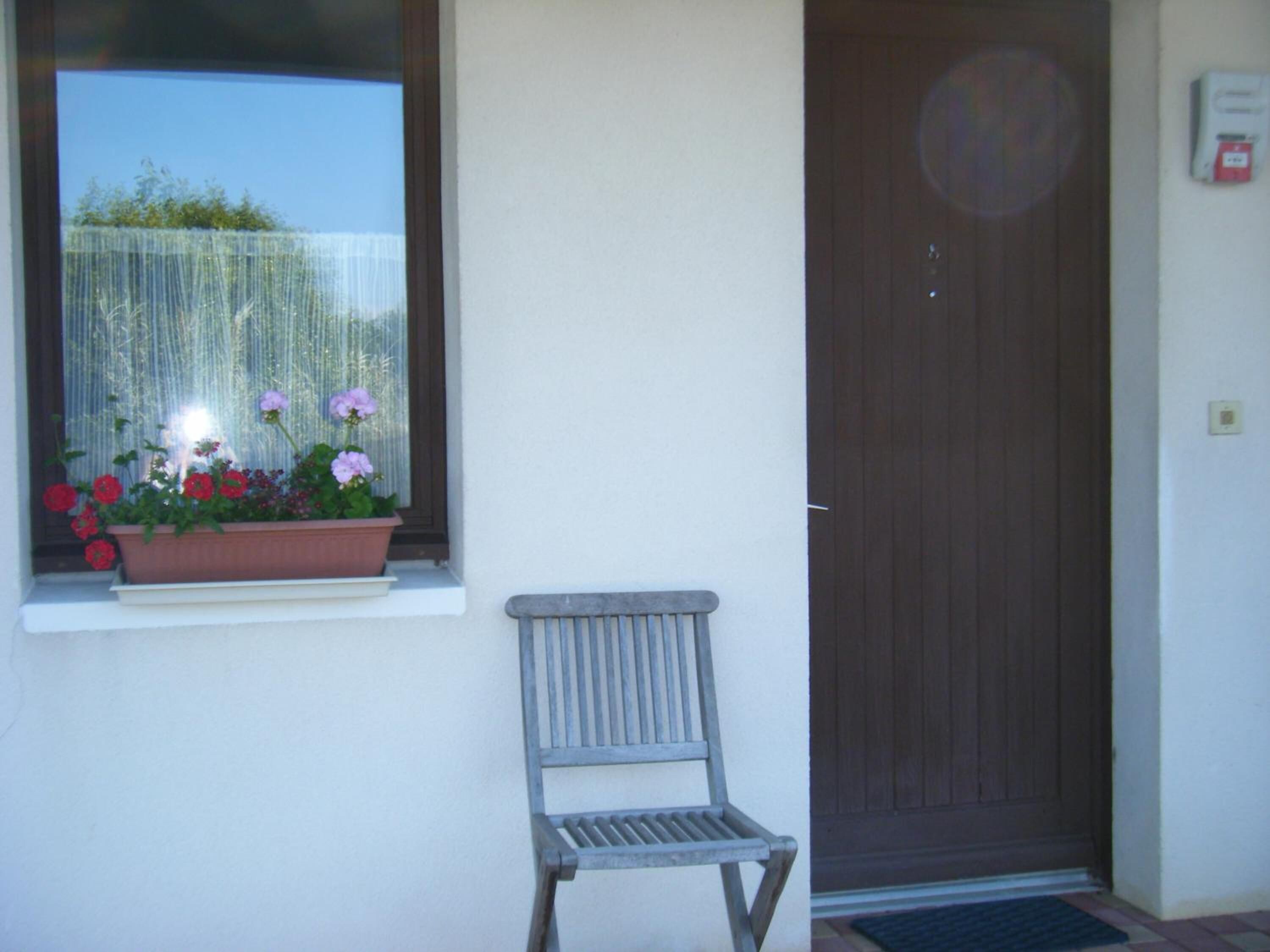 Ein Fenster mit Blumenkasten, der rote und pinke Blumen enthält, ein hölzerner Stuhl und eine braune Tür.