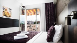 Una habitación de hotel con cama, ventana con cortinas, un cuadro en la pared y un balcón con mesa y sillas.
