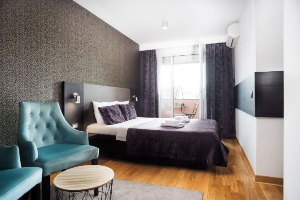 A modern hotel room with a large bed, a tufted armchair, a small round table, and a view of the outdoors through a large window.