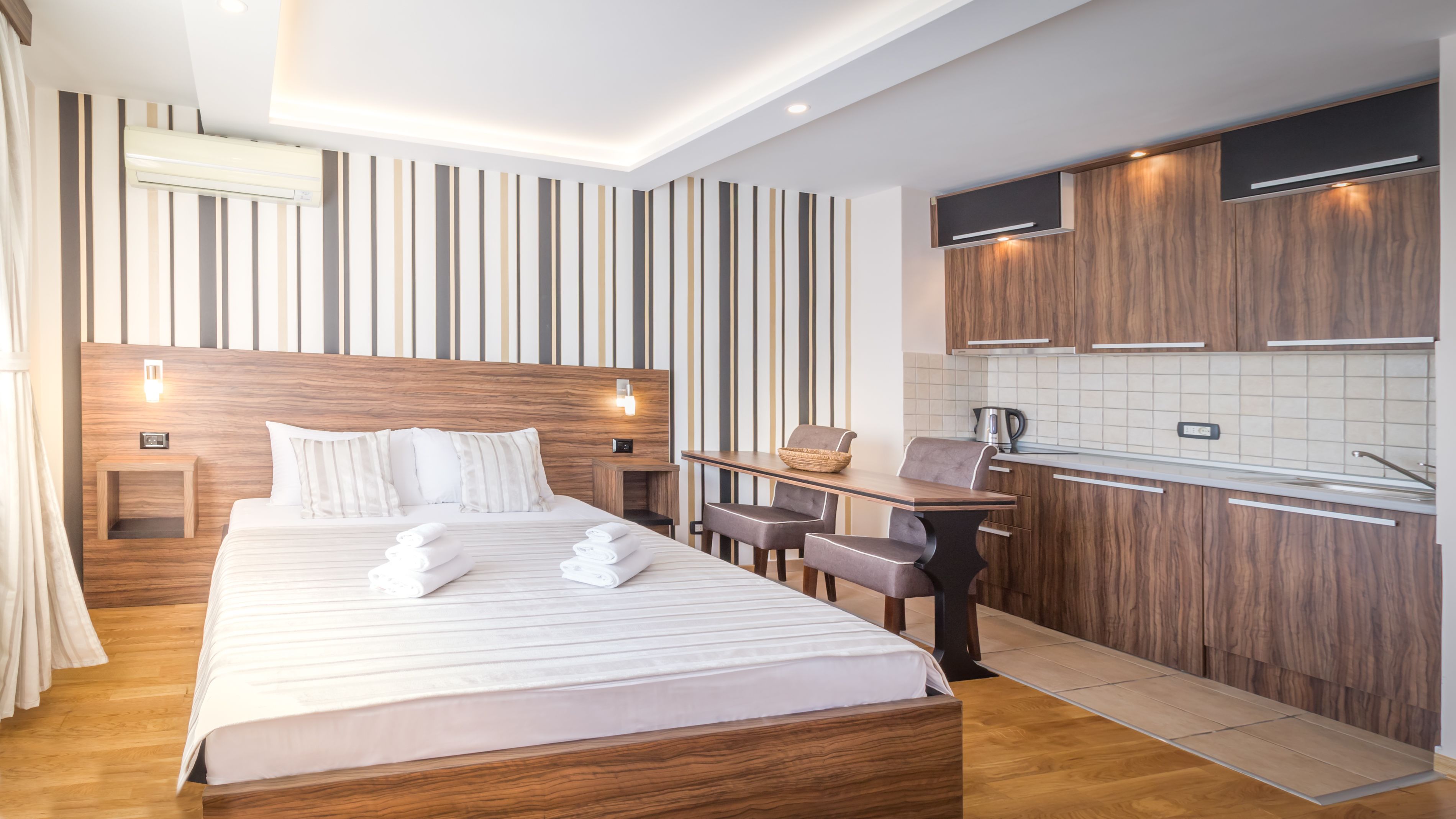 A modern hotel room with a wooden headboard, a large bed with white linens, a small table with chairs, a kitchenette with wooden cabinets, and a striped wall design.