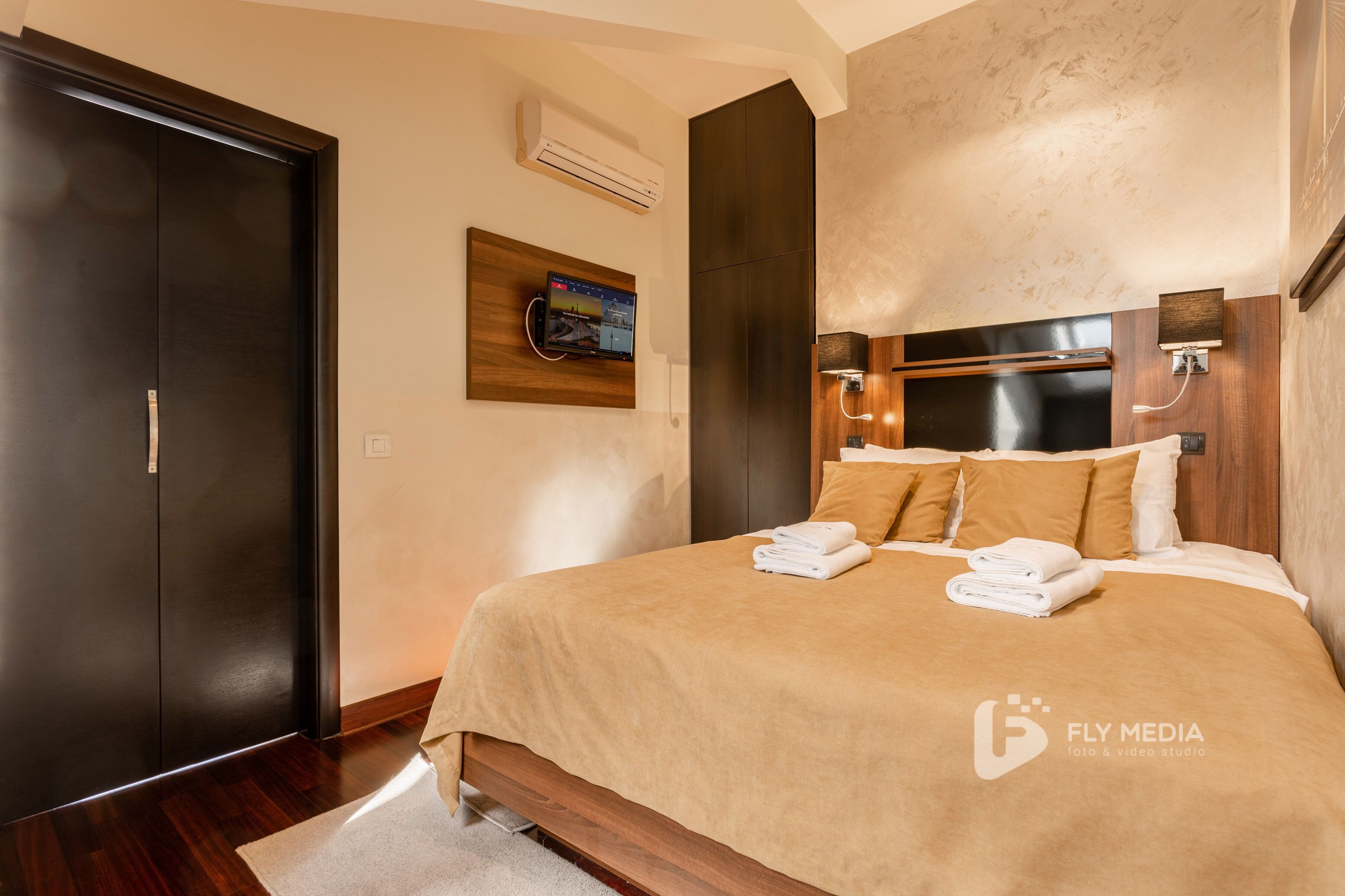 A modern hotel room with a large bed, a flat-screen TV mounted on the wall, and a wooden wardrobe.