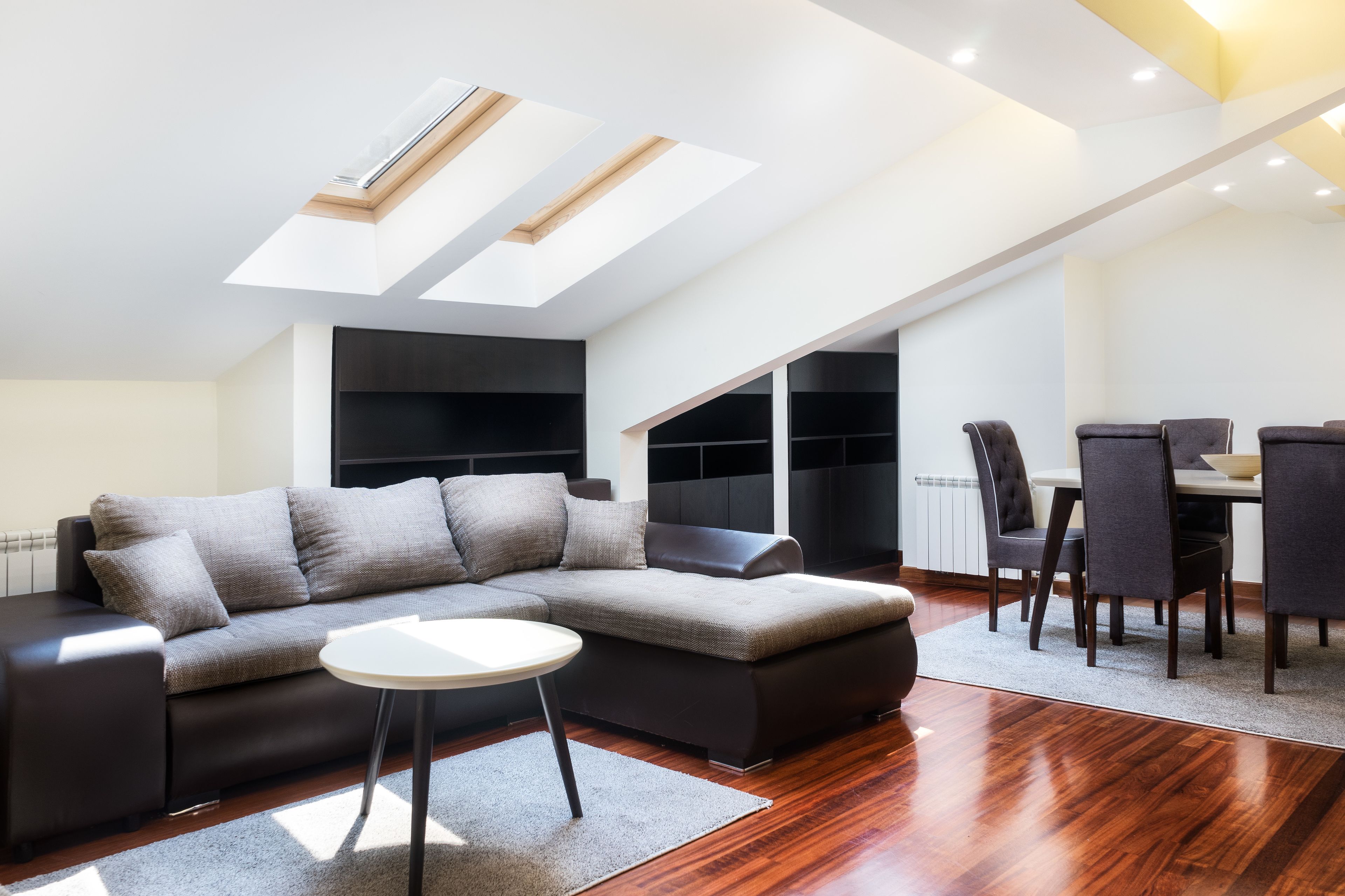 A modern living room with a sectional sofa, a dining table with chairs, and skylights.
