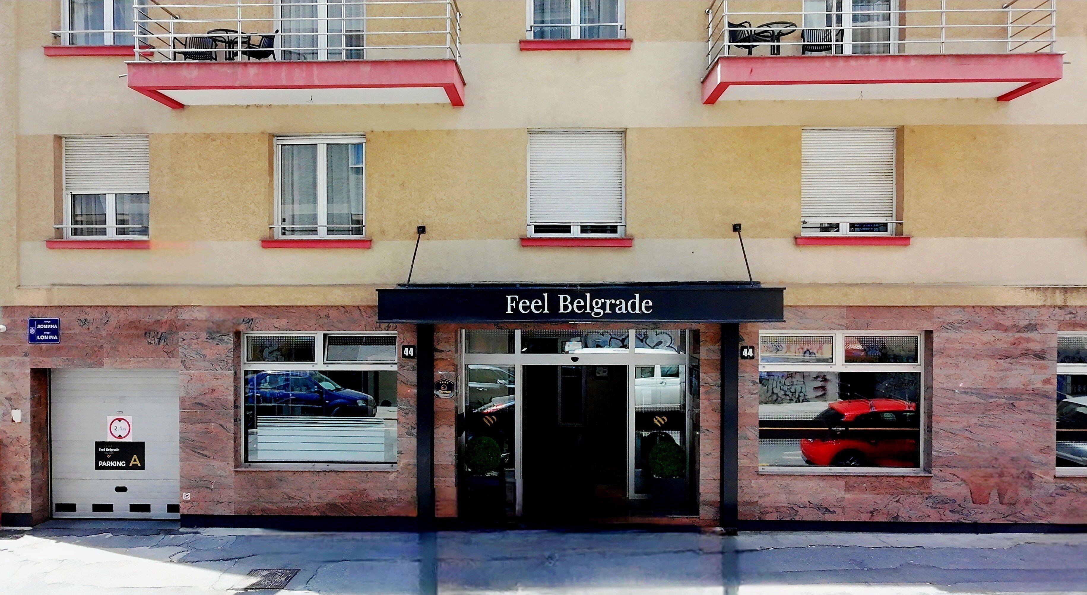 A building with a sign reading "Feel Belgrade" above the entrance. There are windows with blinds, a parking meter, and a fire hydrant visible.