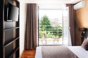 A bedroom with a bed, a TV mounted on the wall, a balcony with chairs, and an air conditioning unit.