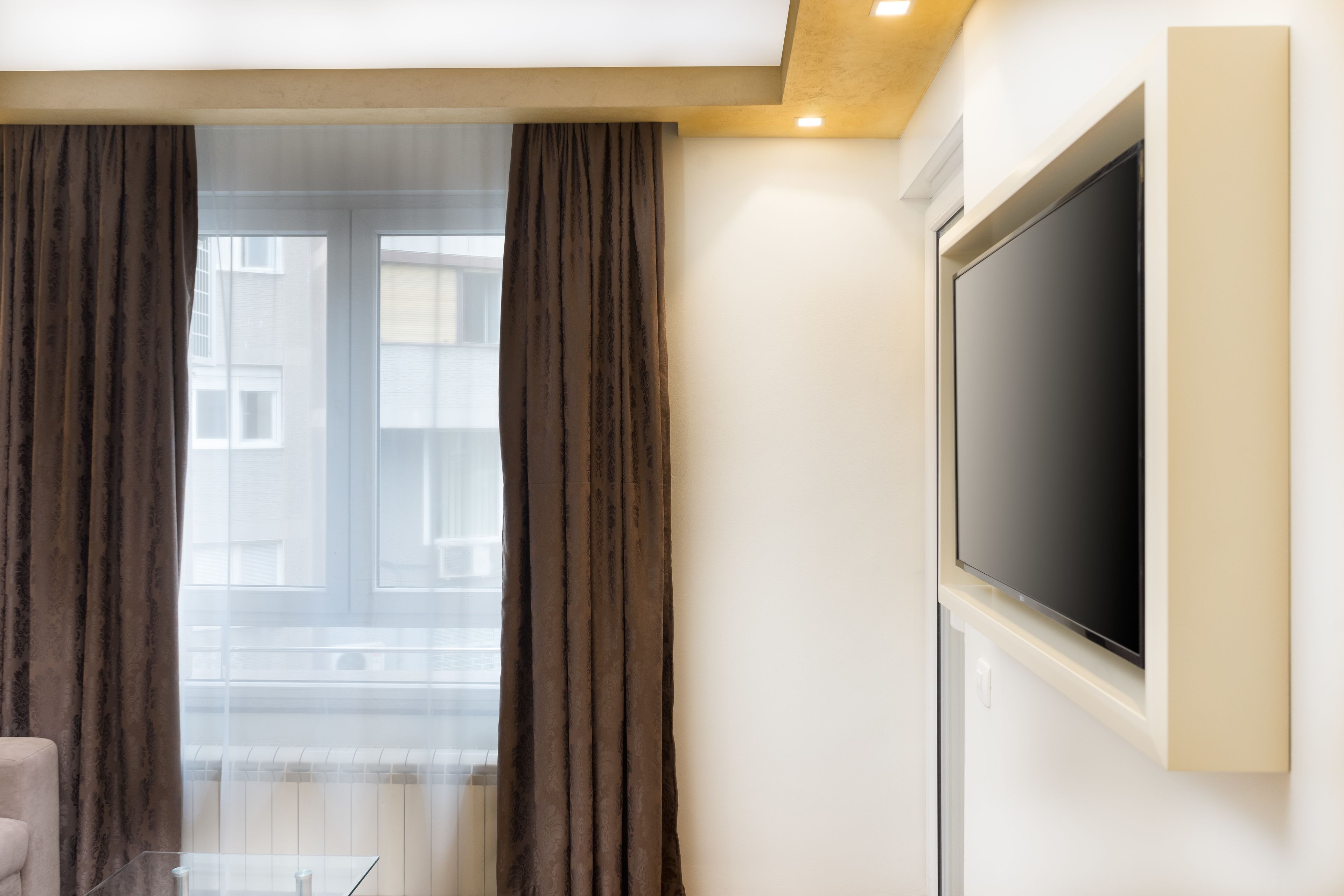 A modern hotel room with a large flat-screen TV mounted on the wall, a large window with curtains, and a clear glass table.