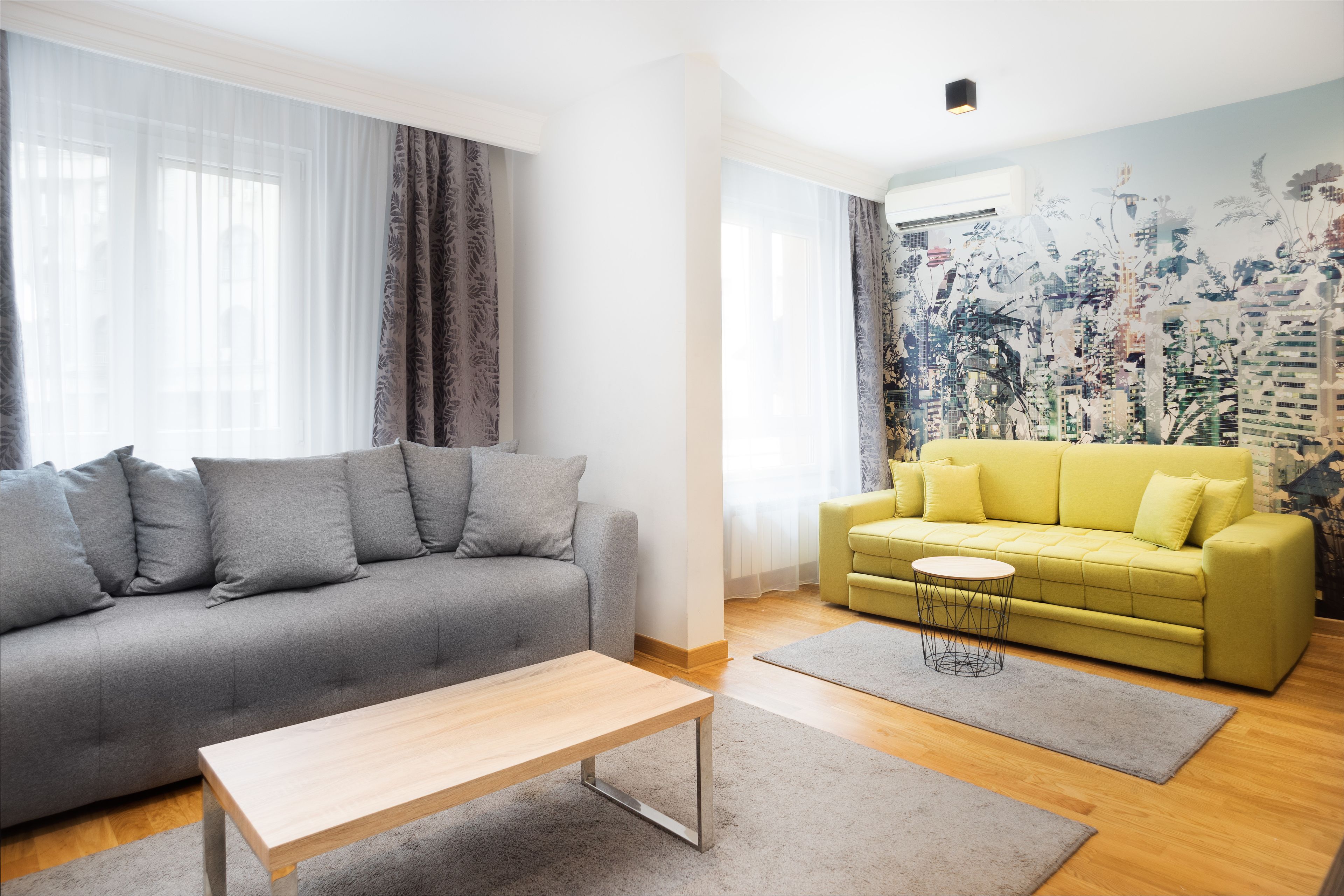 A living room with a grey sofa, a yellow sofa, a wooden coffee table, and a cityscape mural on the wall.