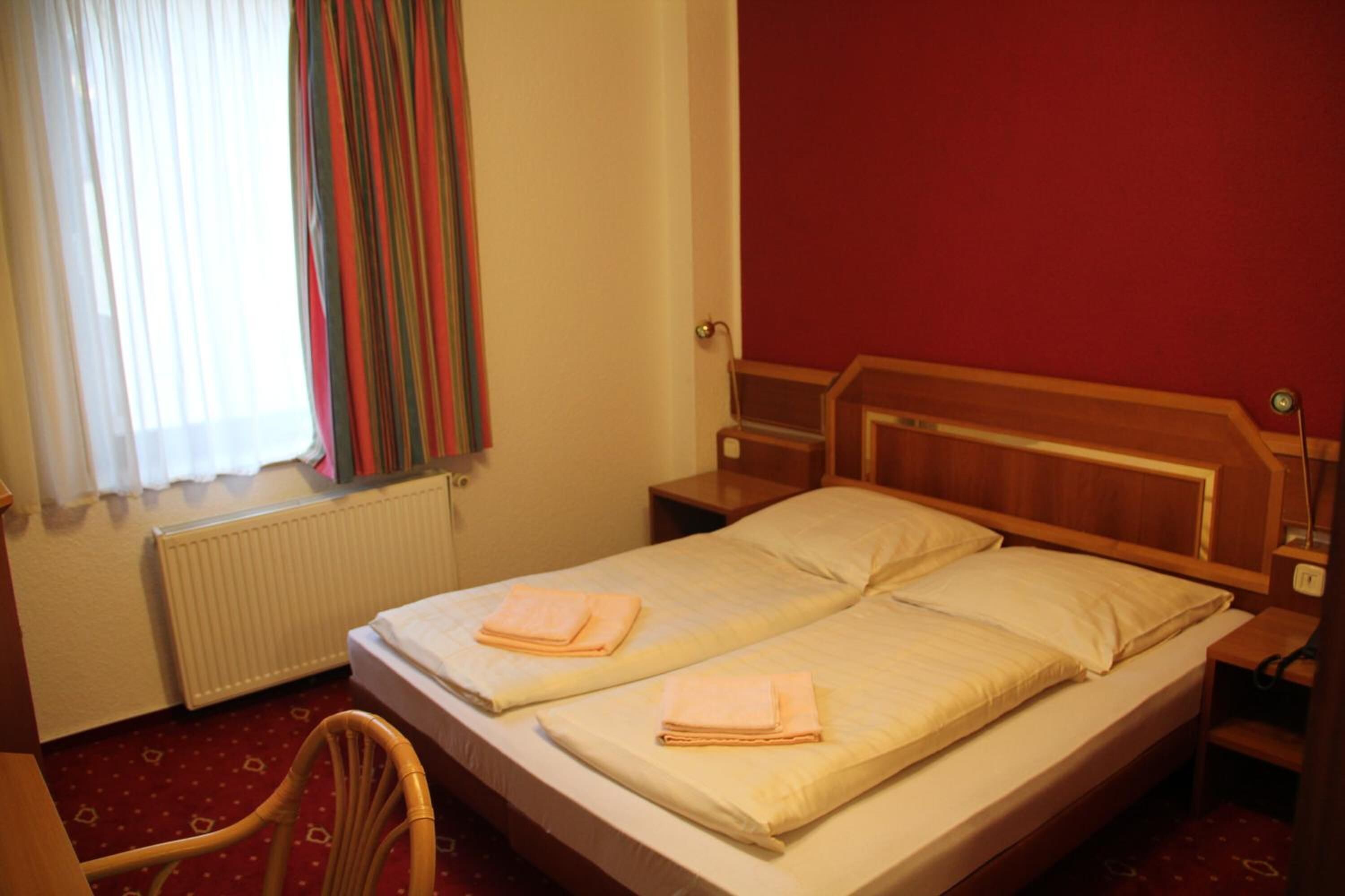 A double bed with two pillows and folded towels, a wooden headboard, a nightstand, a chair, a window with curtains, and a radiator.