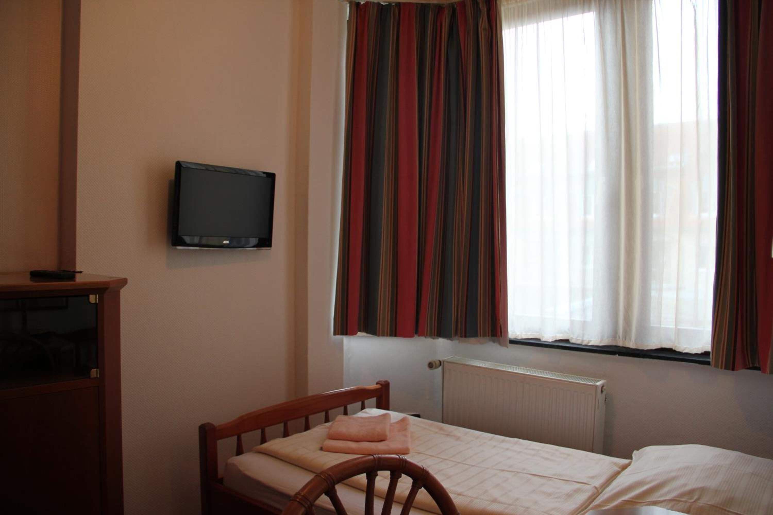 A small, single-bed room with a wooden headboard, a television mounted on the wall, and a window with striped curtains.