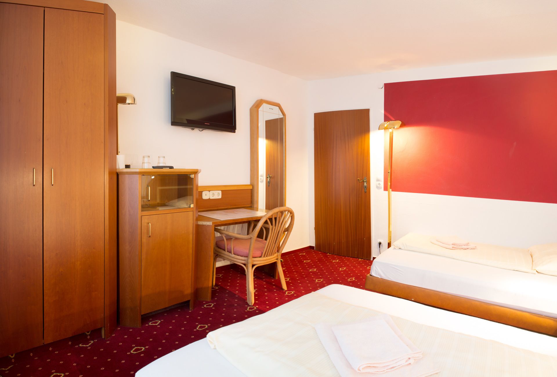 A hotel room with a bed, a desk with a chair, a wardrobe, a television, and a mirror.