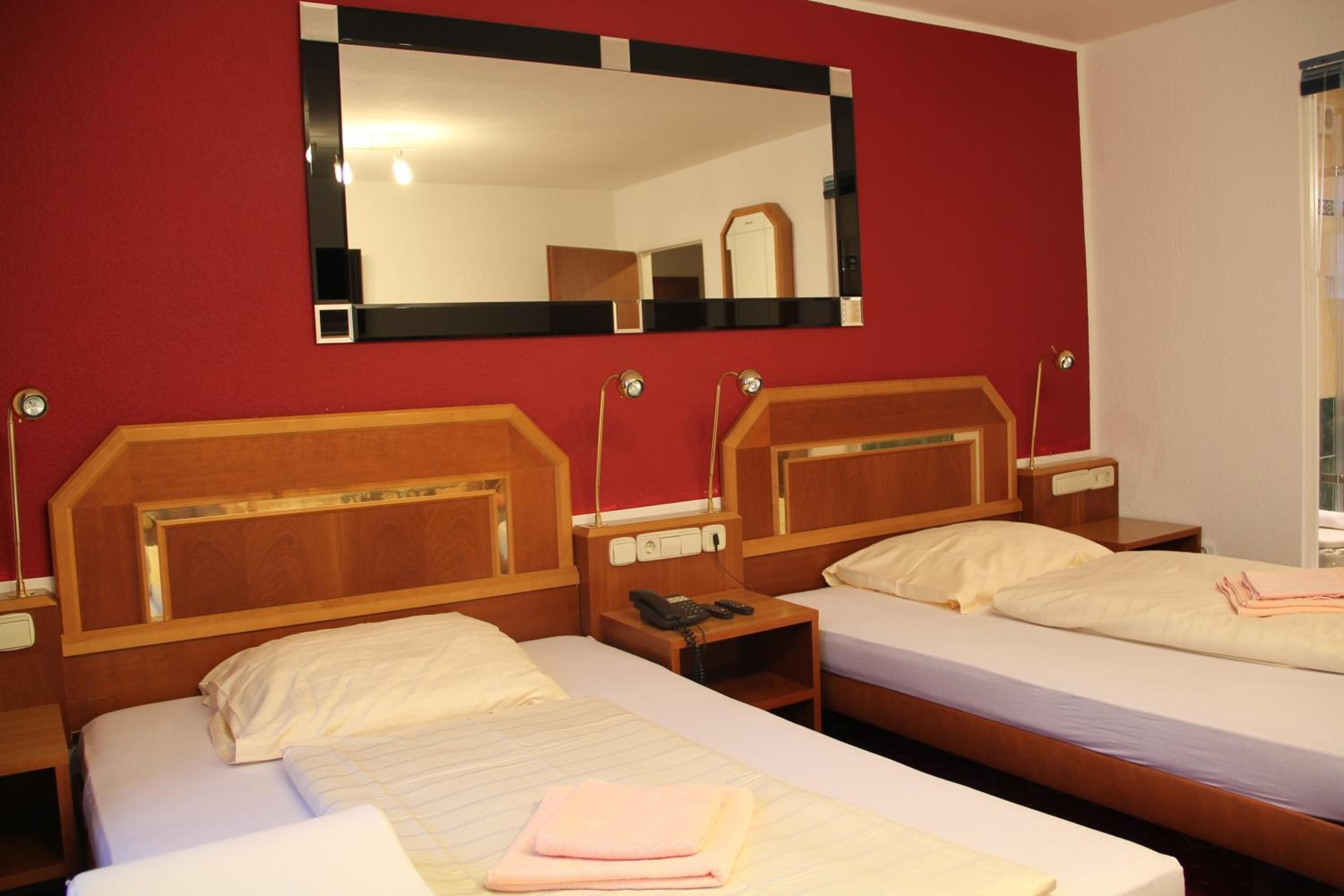 A hotel room with two single beds, wooden headboards, bedside tables with lamps, and a large mirror on the wall.