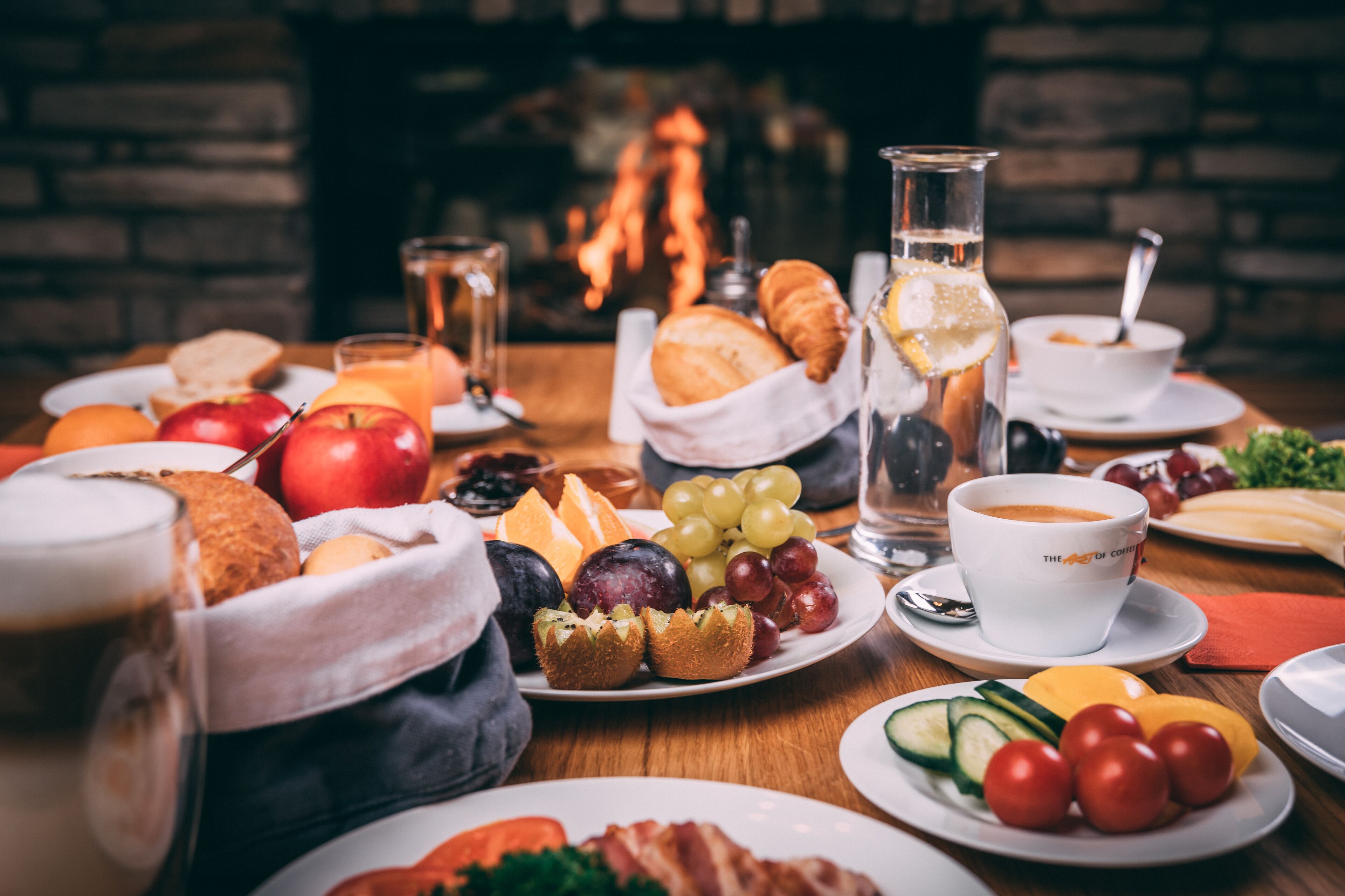 A breakfast setting with various foods and drinks on a wooden table, a fireplace in the background, and a cozy atmosphere.