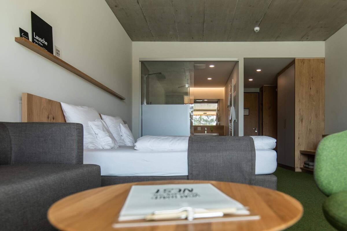 Una habitación de hotel moderna con una cama, un sofá gris, una mesa de centro de madera y vistas al baño.