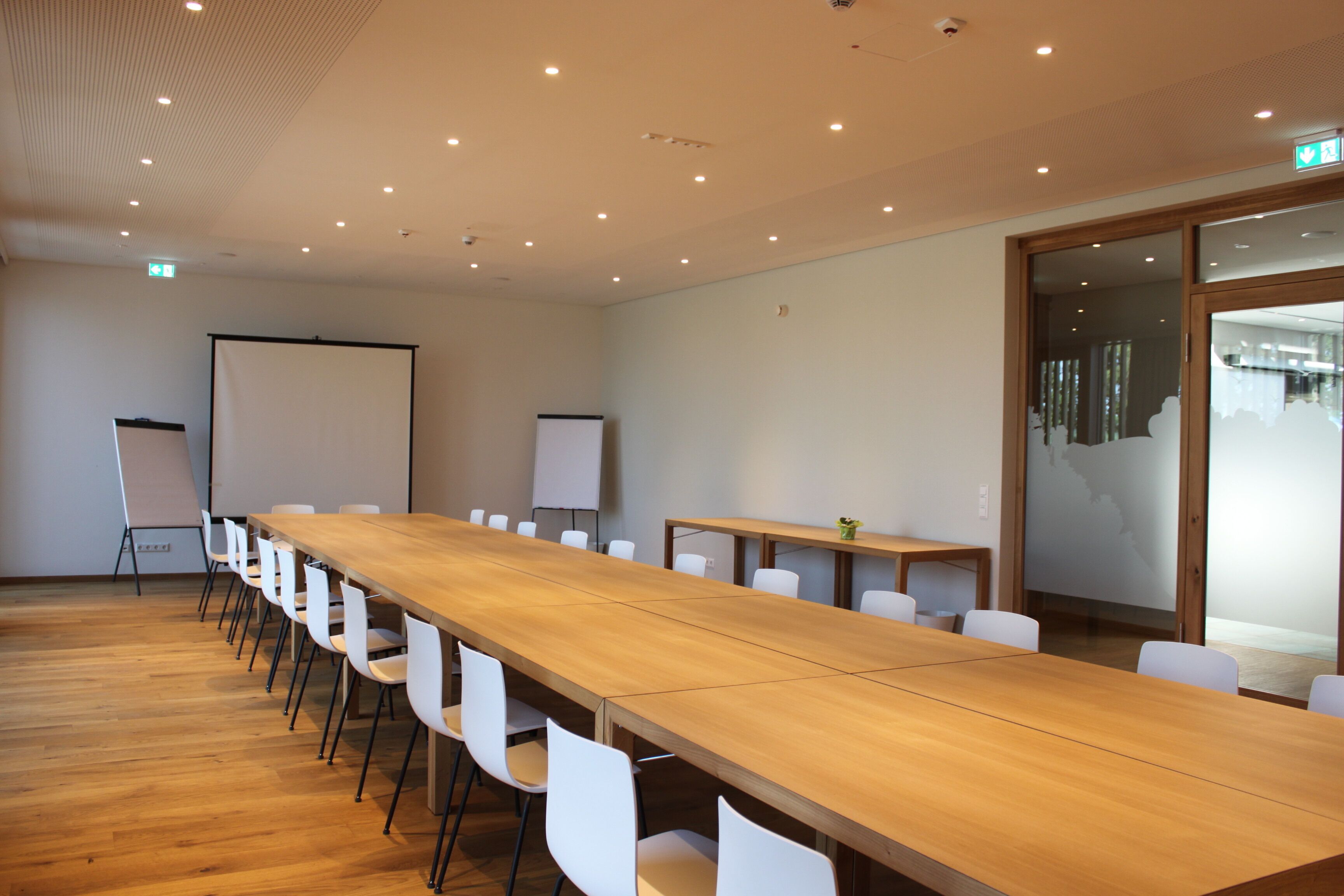 A conference room with a long wooden table, white chairs, a whiteboard, and a projection screen.
