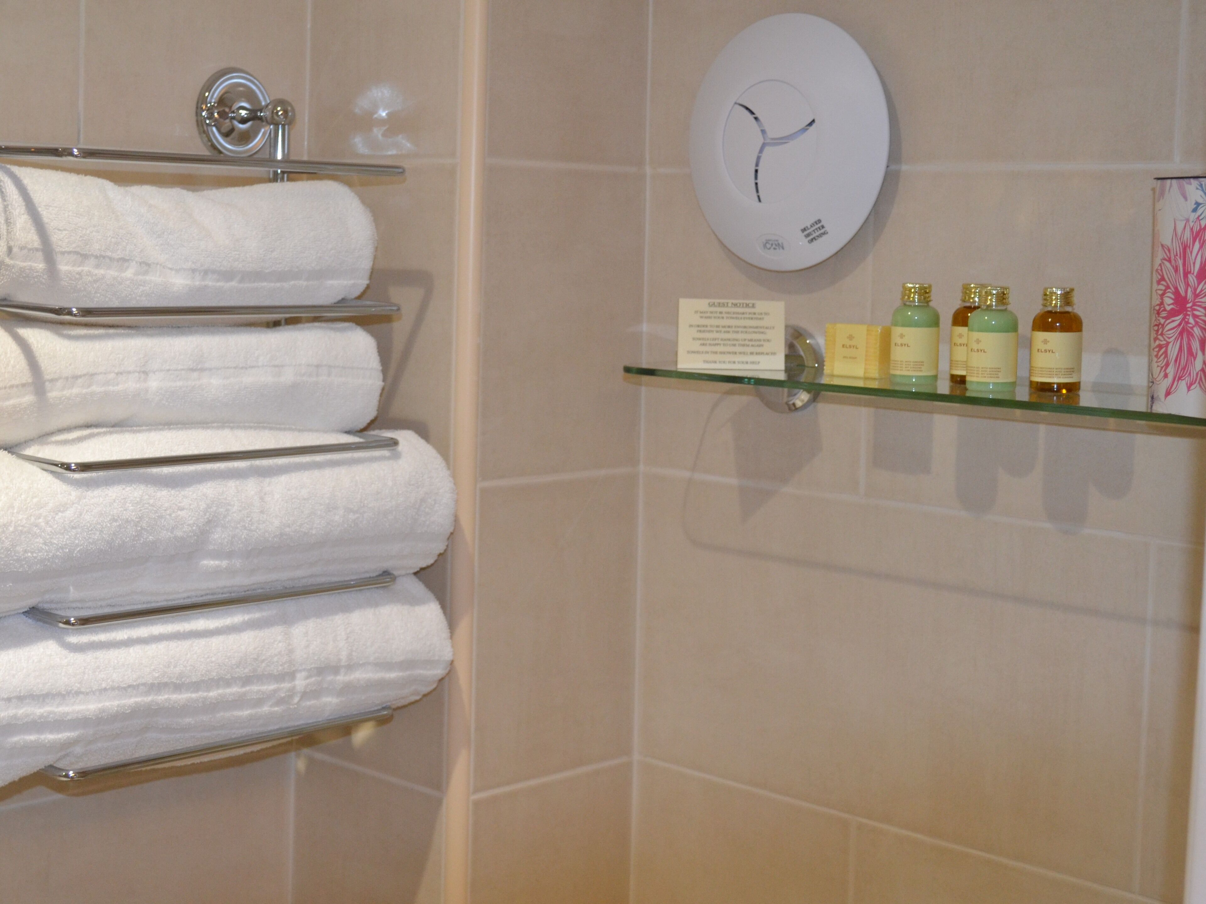Un baño con un estante de vidrio que sostiene artículos de aseo, un perchero con toallas dobladas y un reloj de pared.