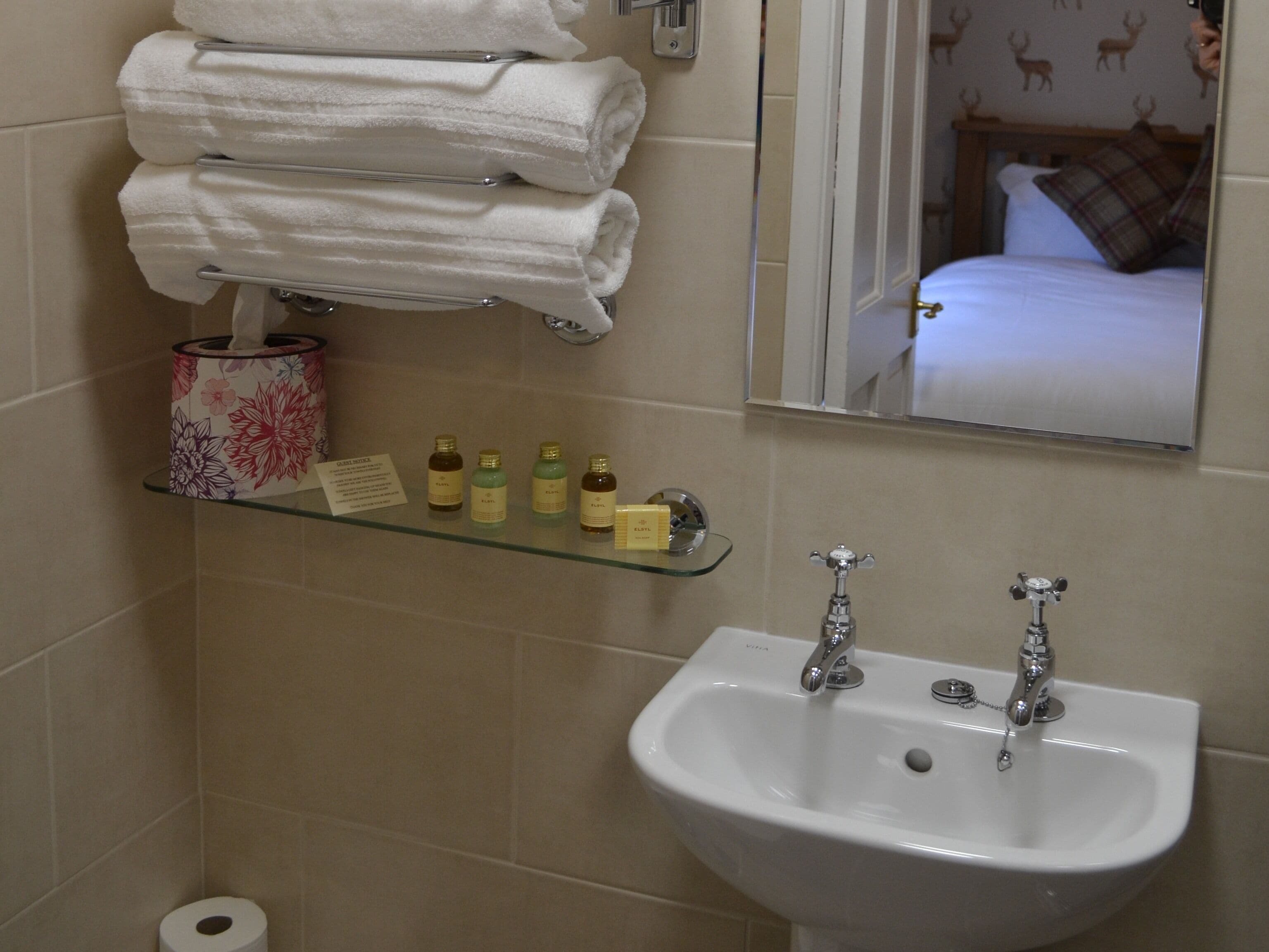 Un baño con un lavamanos, dos grifos, un estante de vidrio con artículos de tocador, un perchero con toallas dobladas y un dispensador de papel higiénico.