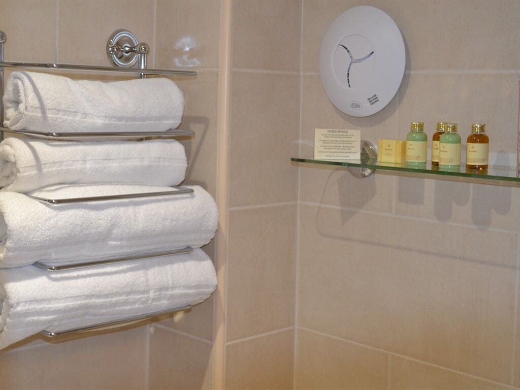A bathroom with a towel rack holding white towels, a glass shelf with toiletries, and a wall clock.