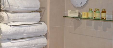 A bathroom with a towel rack holding white towels, a glass shelf with toiletries, and a wall clock.