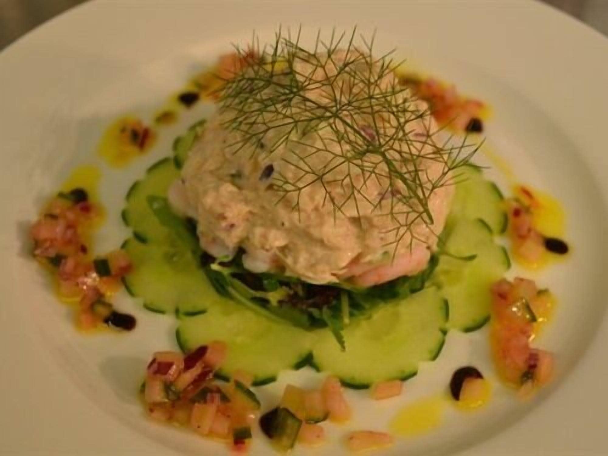 A plate with a layered dish, possibly a salad or appetizer, topped with dill and surrounded by cucumber slices and a small amount of dressing.