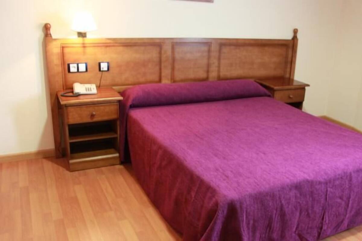 Ein Schlafzimmer mit einem Holz-Kopfteil, einer violetten Bettdecke, einem Nachttisch mit Lampe und einer kleinen Schublade.