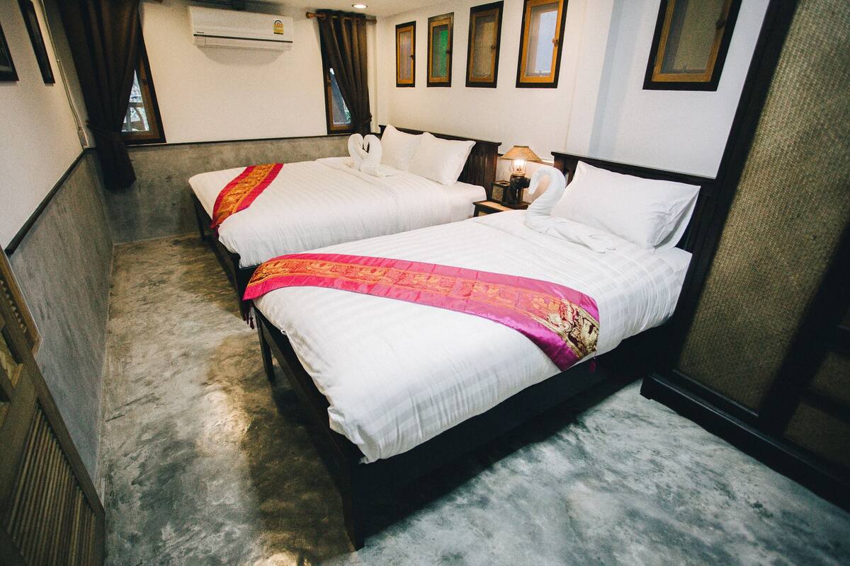 A hotel room with two beds, each with white bedding and a red and gold decorative headboard. There is a nightstand with a lamp and a wall-mounted air conditioner.