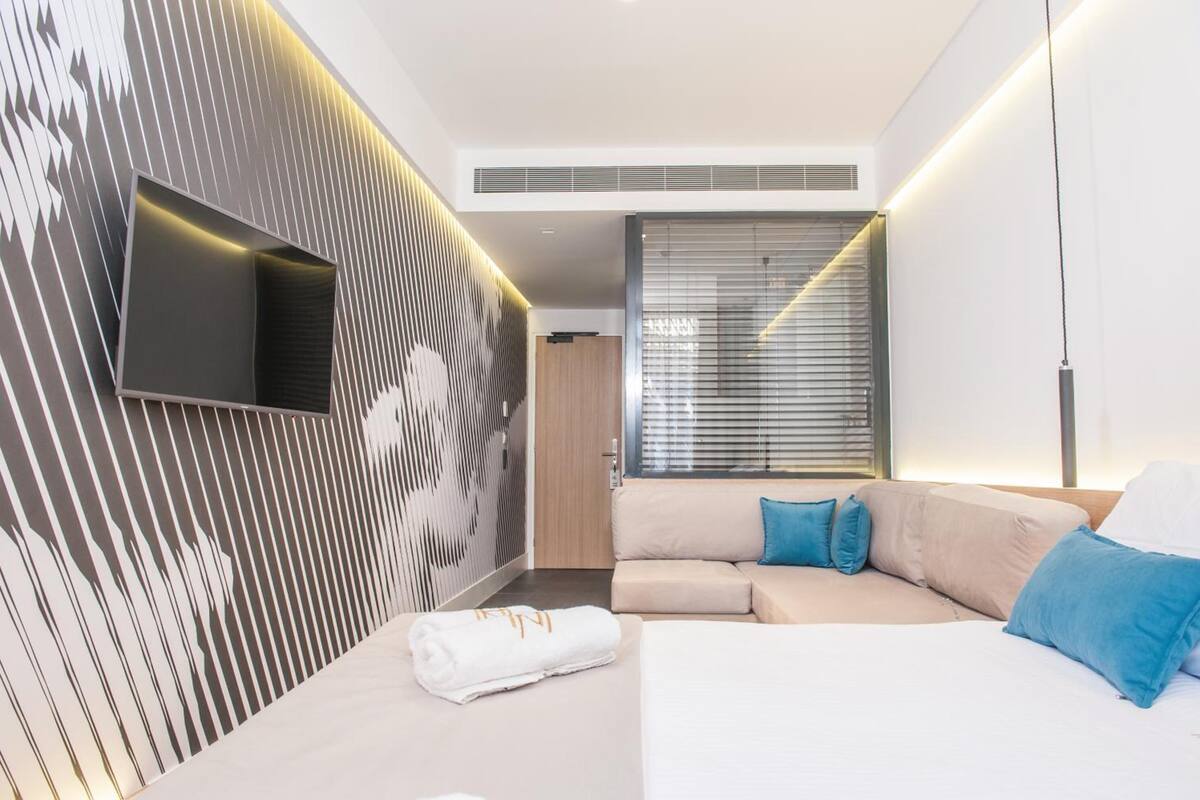 A modern hotel room with a flat-screen TV mounted on a textured wall, a sofa with blue cushions, a neatly made bed, and a large window with blinds.