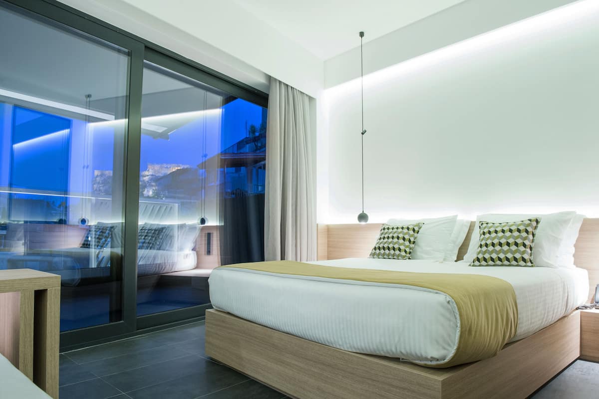 A modern bedroom with a large bed, a glass door leading to a balcony, and a view of the city at dusk.