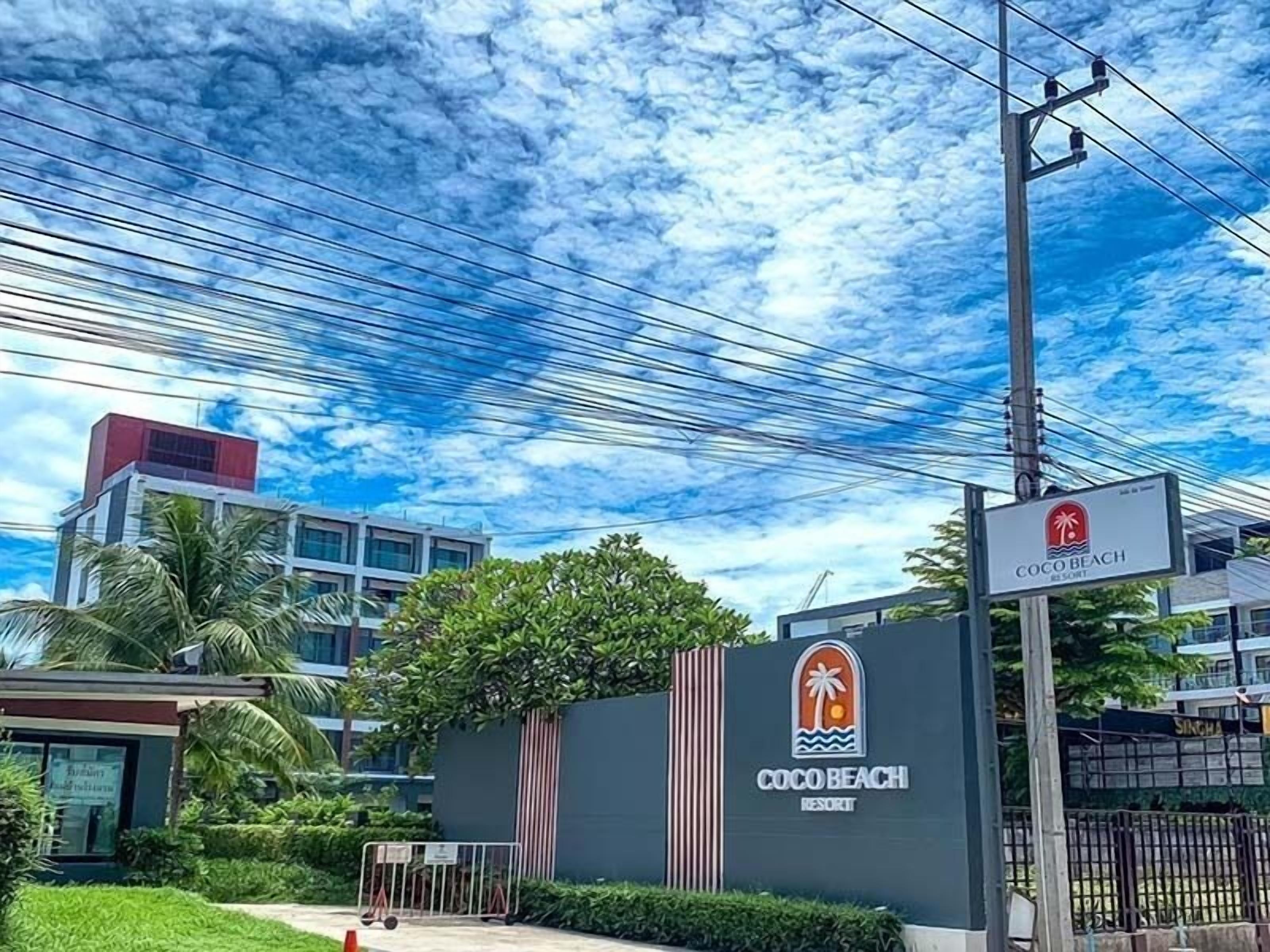 Letrero del Resort Coco Beach con logotipo de palmera, rodeado de follaje y edificios modernos.