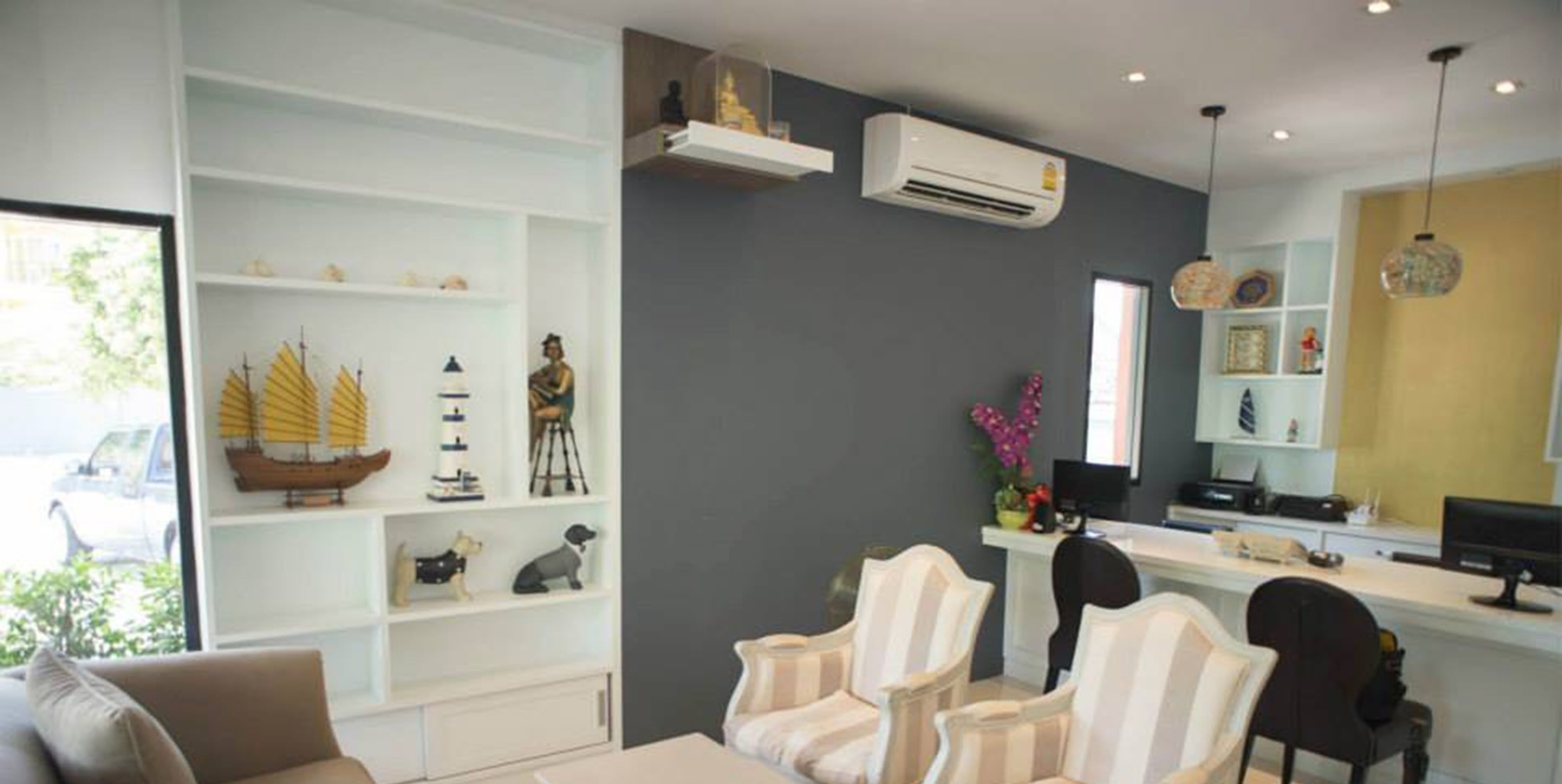 Una sala de estar moderna con un sofá blanco, sillones a rayas y un escritorio con una computadora. Se observan estantes con objetos decorativos y un equipo de aire acondicionado.
