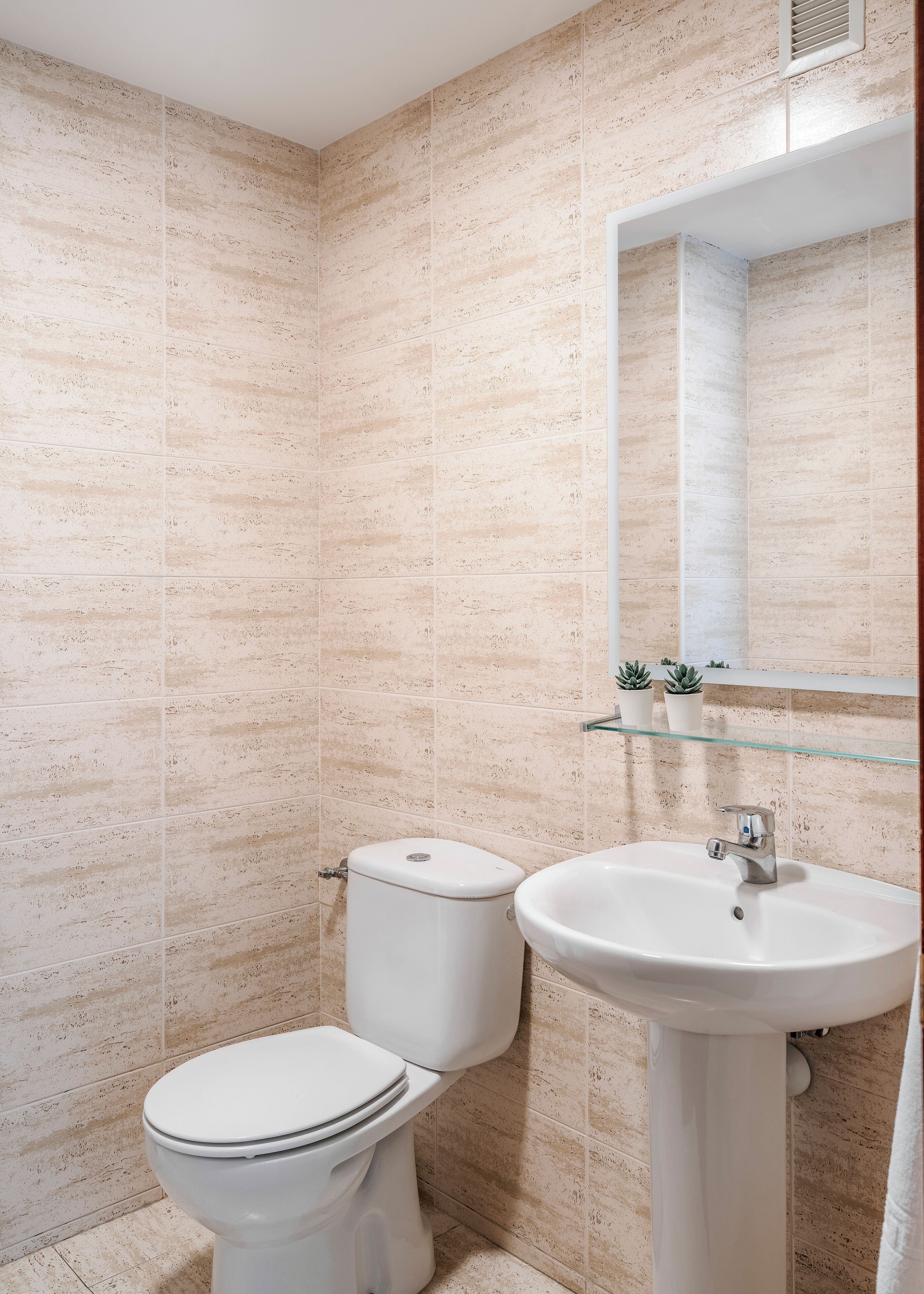 A bathroom with beige tiled walls, a white toilet, and a white sink with a glass shelf above it.