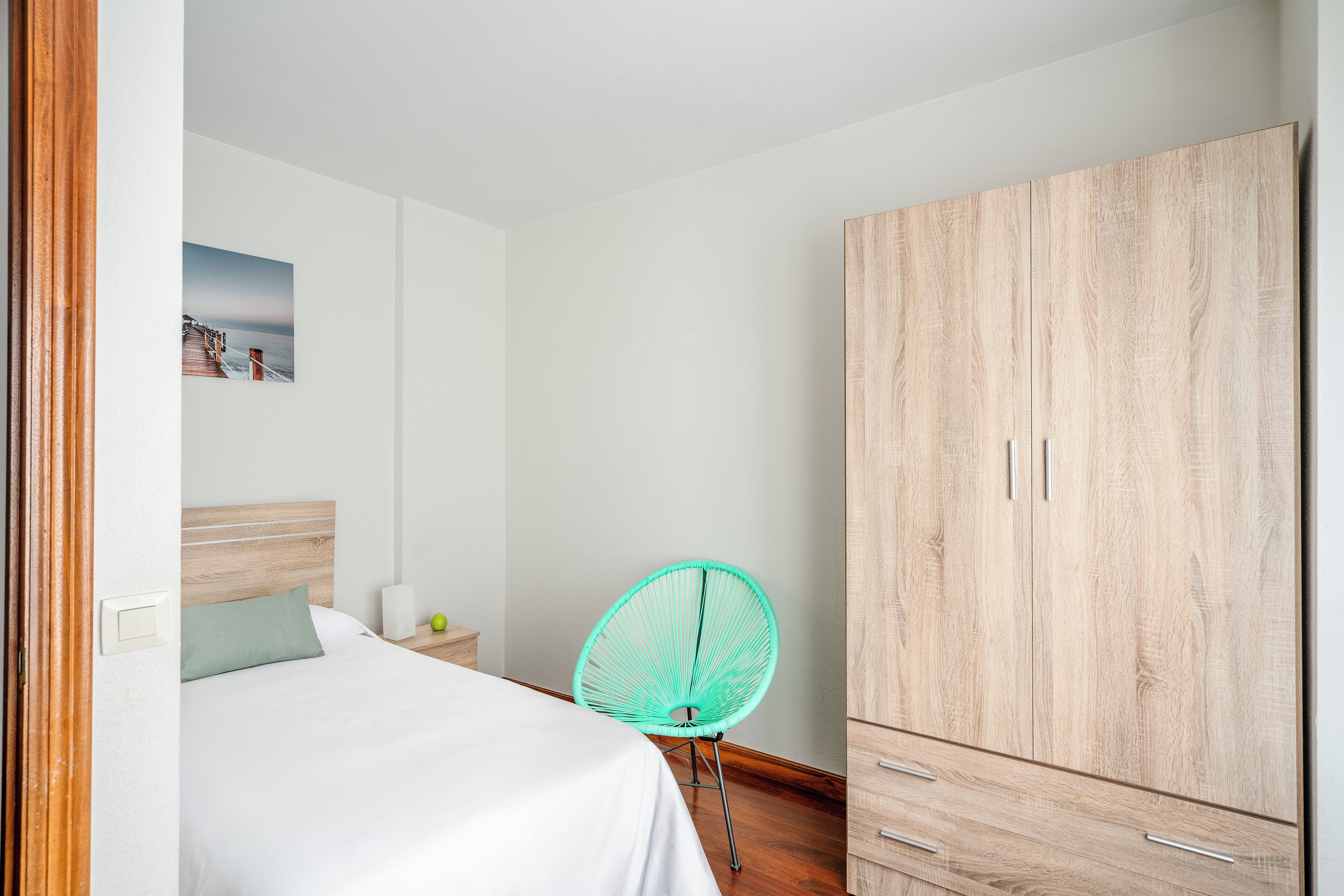 A bedroom with a bed, a wooden wardrobe, a green chair, and a wall-mounted picture.