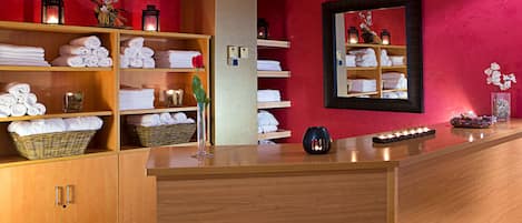 A hotel reception area with wooden cabinets, shelves with folded towels, a vase with flowers, and a mirror on the wall.