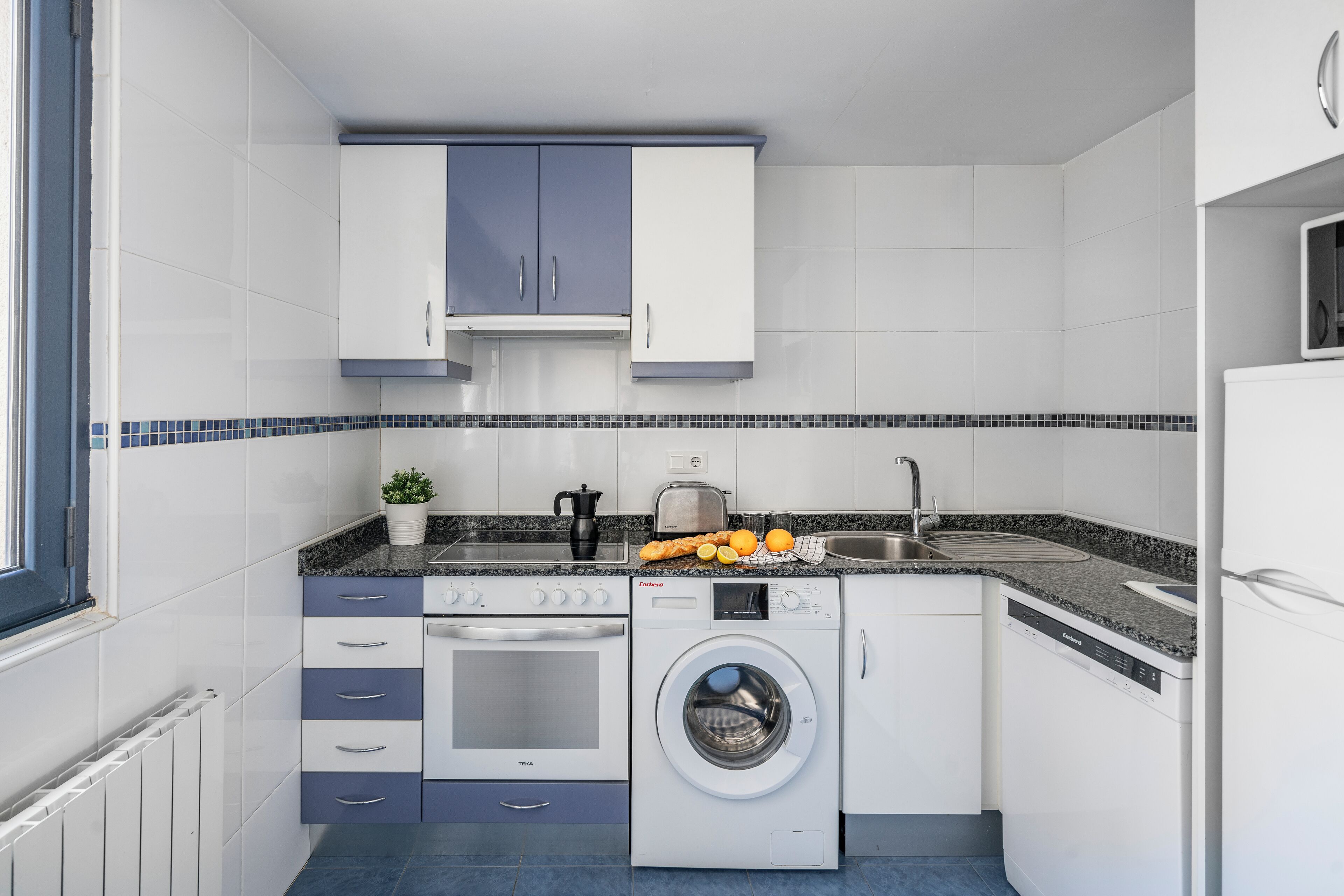 A compact kitchen with white and blue cabinets, a built-in oven, a washing machine, and a countertop with a kettle and oranges.