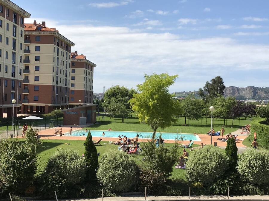 Zona de piscina con gente tomando el sol y nadando, rodeada de vegetación y un edificio de varios pisos.