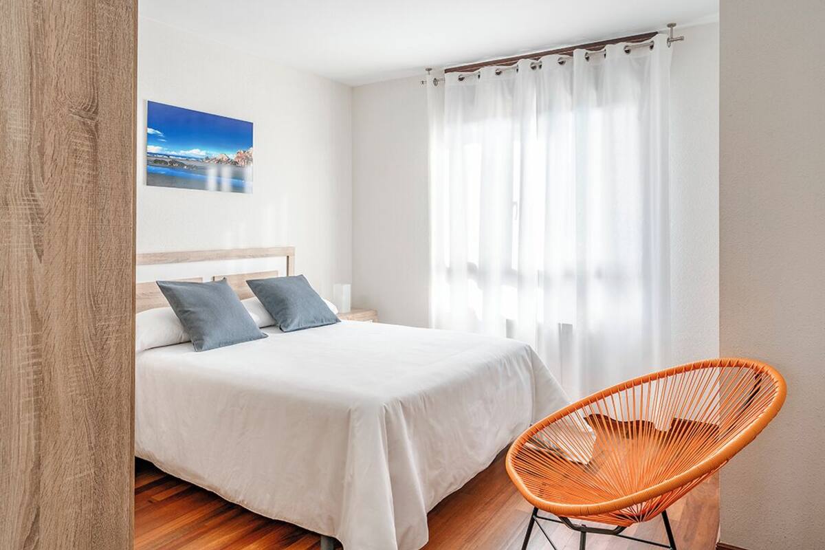 A bedroom with a bed, a chair, a picture on the wall, and a window with curtains.
