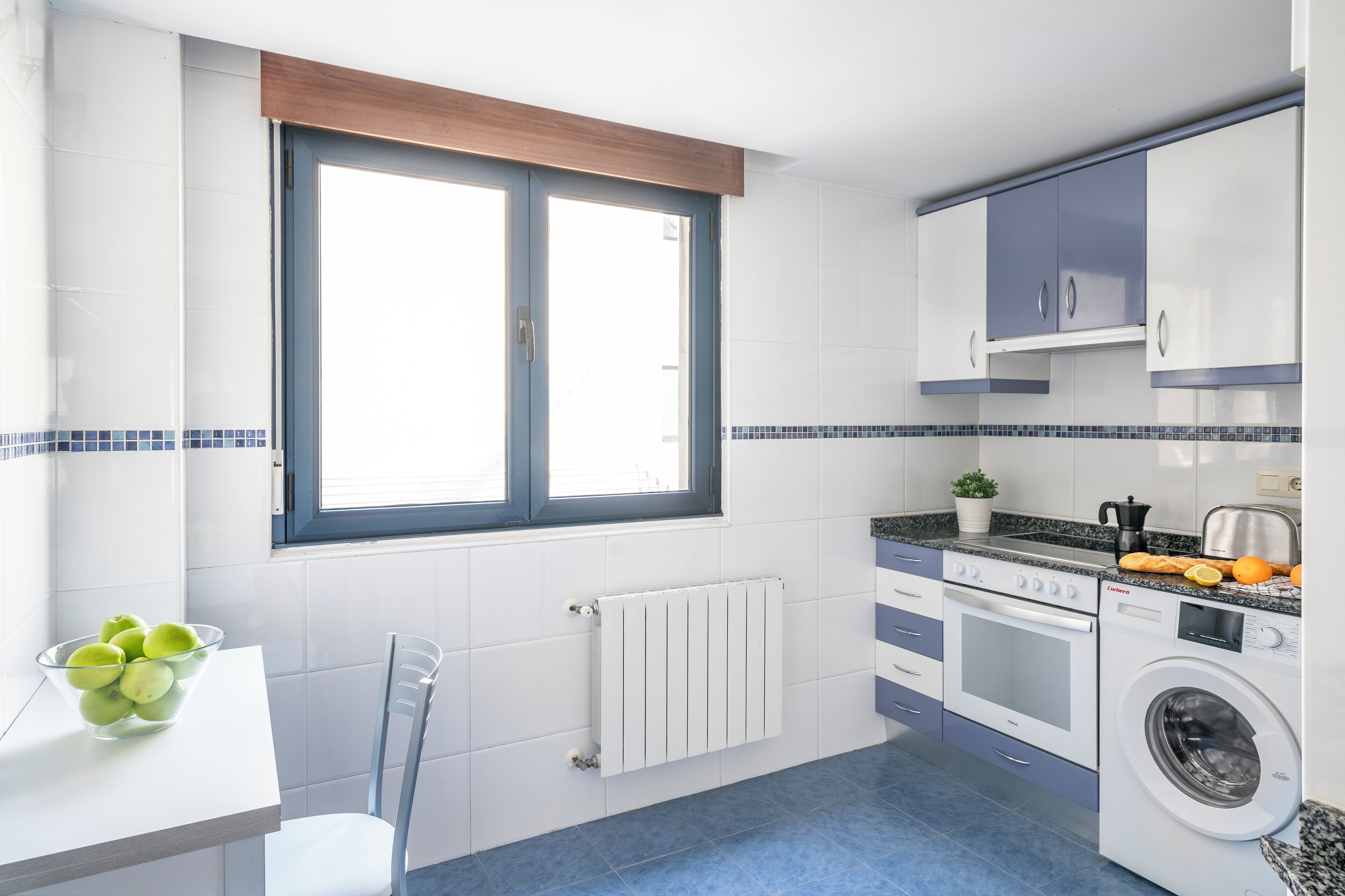 Une cuisine avec une fenêtre, des placards blancs, un lave-linge et un saladier de fruits posé sur le plan de travail.