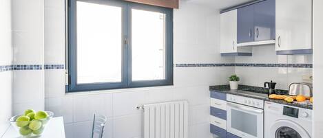 Une cuisine avec une fenêtre, des placards blancs, un lave-linge et un saladier de fruits posé sur le plan de travail.
