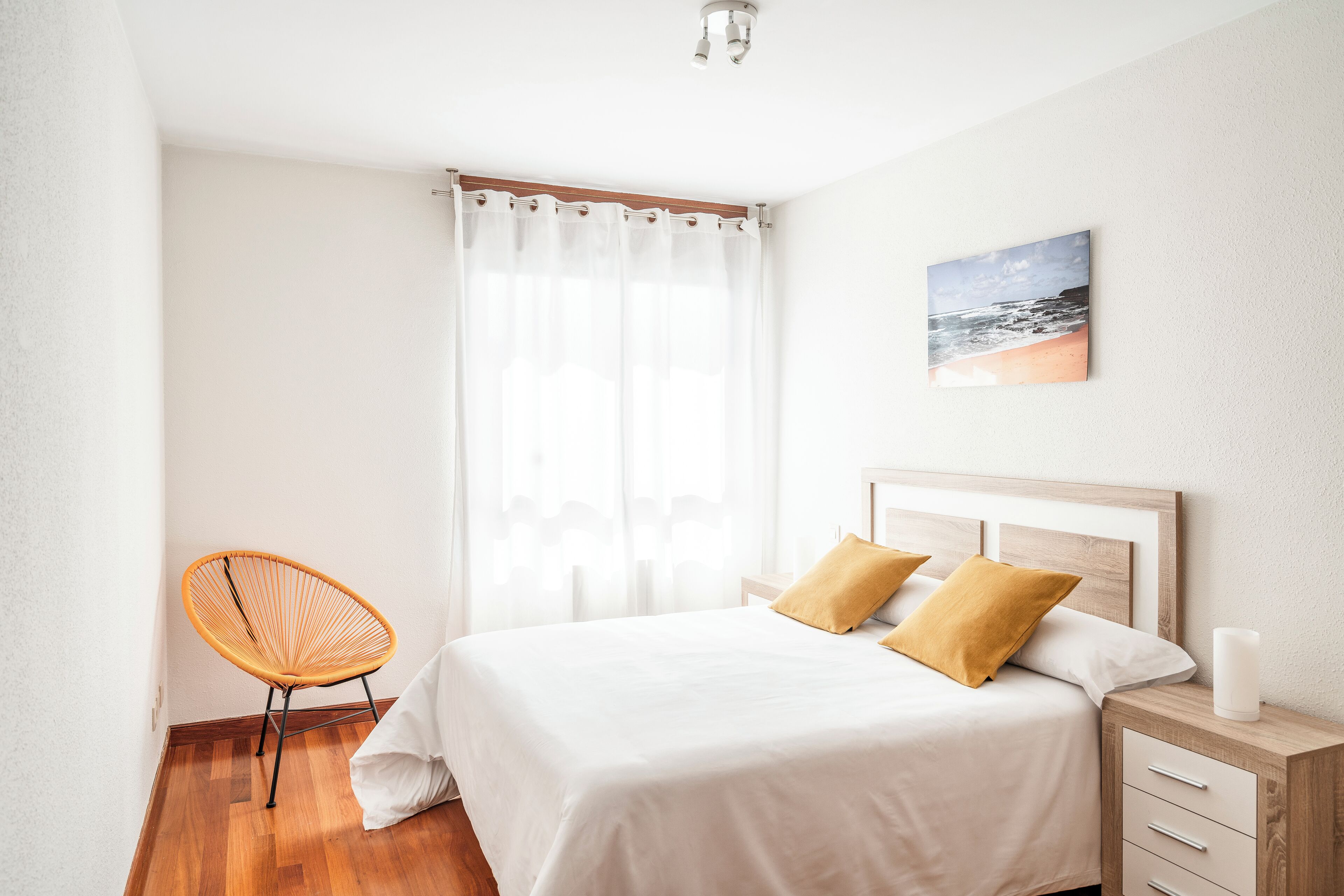 Un dormitorio ordenado con cama, una silla, una mesita de noche y un cuadro en la pared.