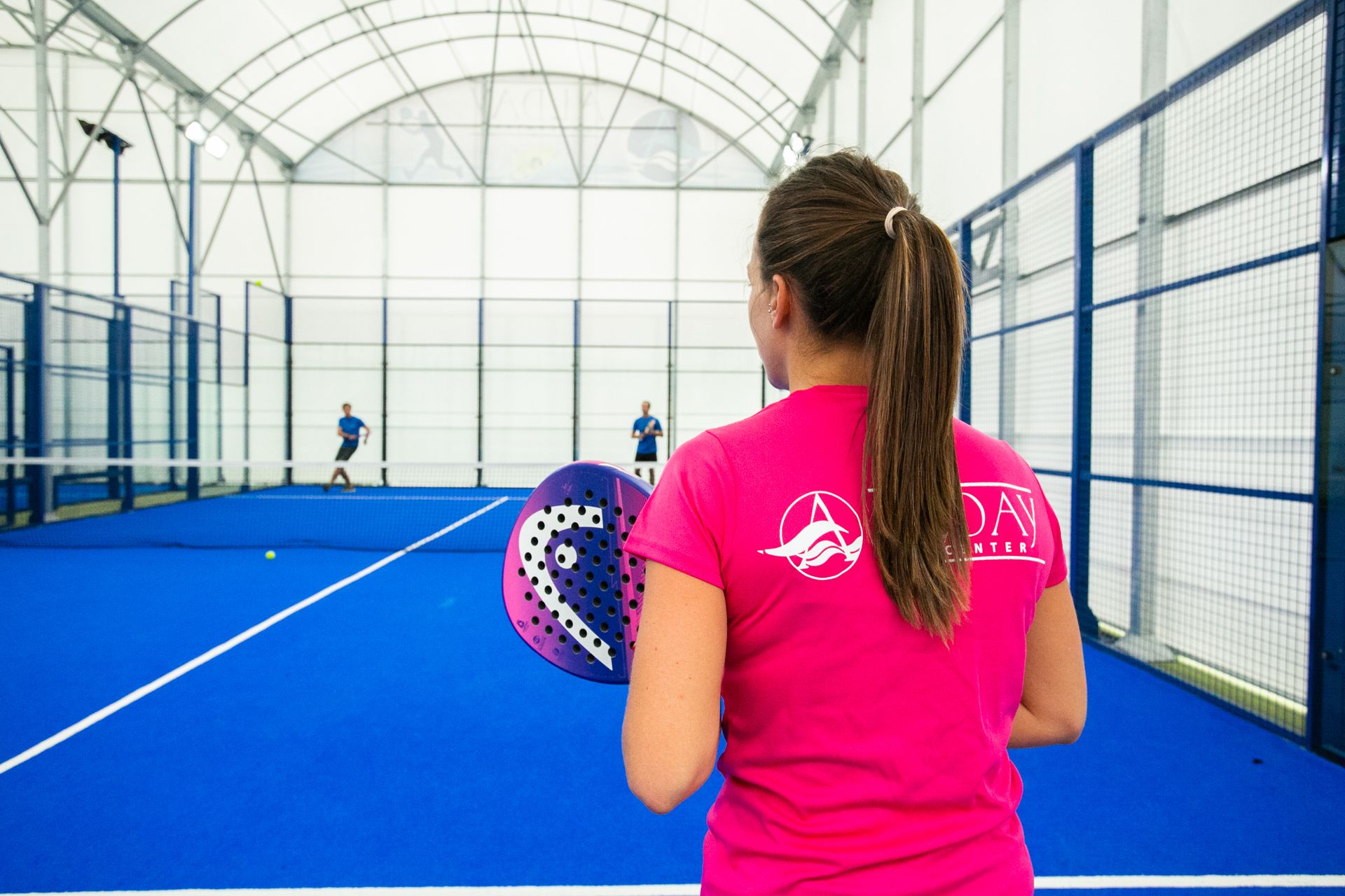 An indoor sports facility with a blue floor, a netted wall, and a person in a pink shirt holding a paddle.